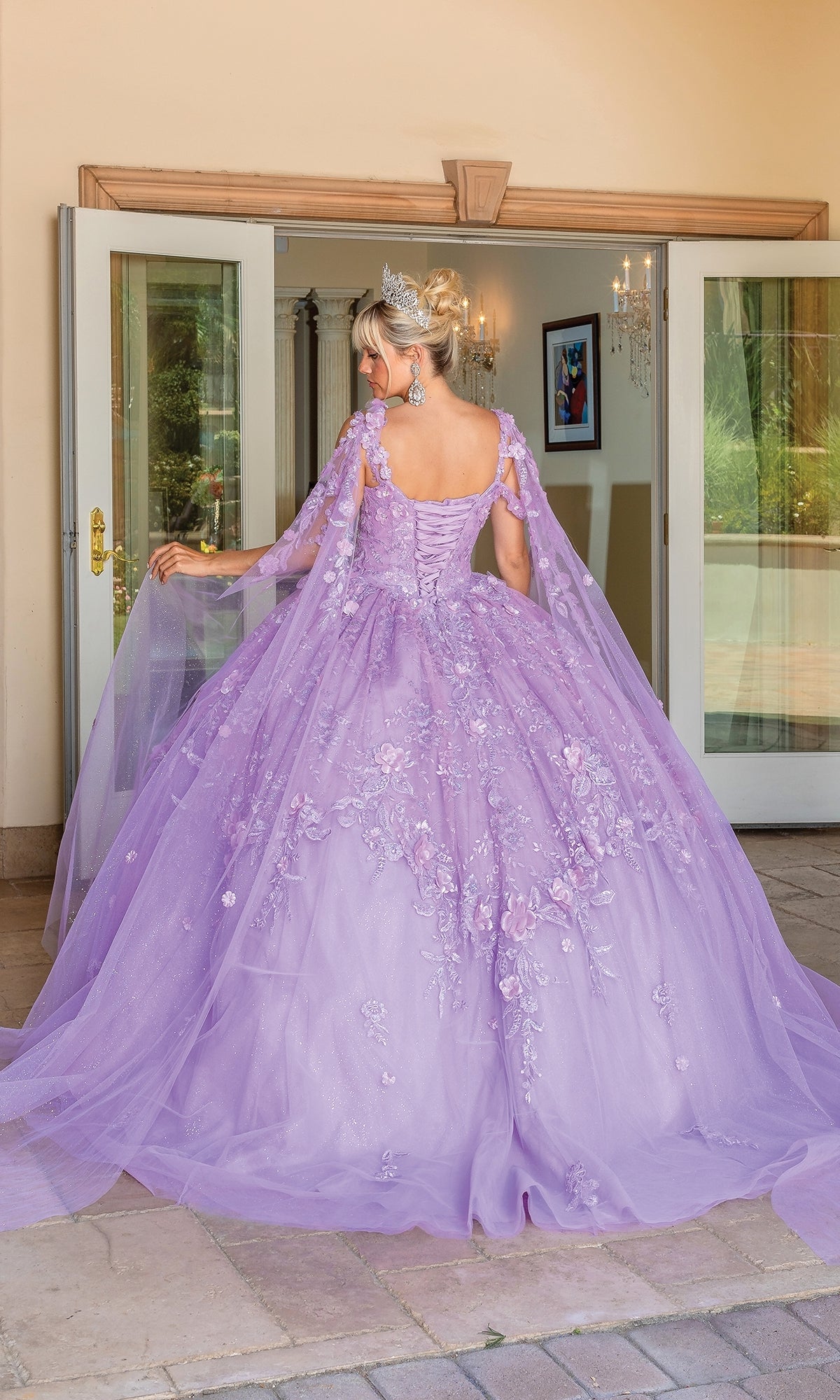 A woman stands before open glass doors, seen from behind, wearing the Quinceanera Dress 1664 By Dancing Queen—a lavish ball gown with lace-embroidered floral details and cape sleeves.