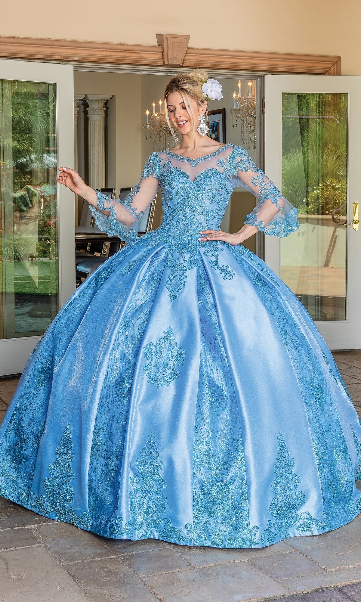 A woman stands outside in front of double doors, wearing the Quinceanera Dress 1665 By Dancing Queen—a light blue ball gown with lace details, sheer bell sleeves, a sweetheart neckline, and a white flower in her hair.