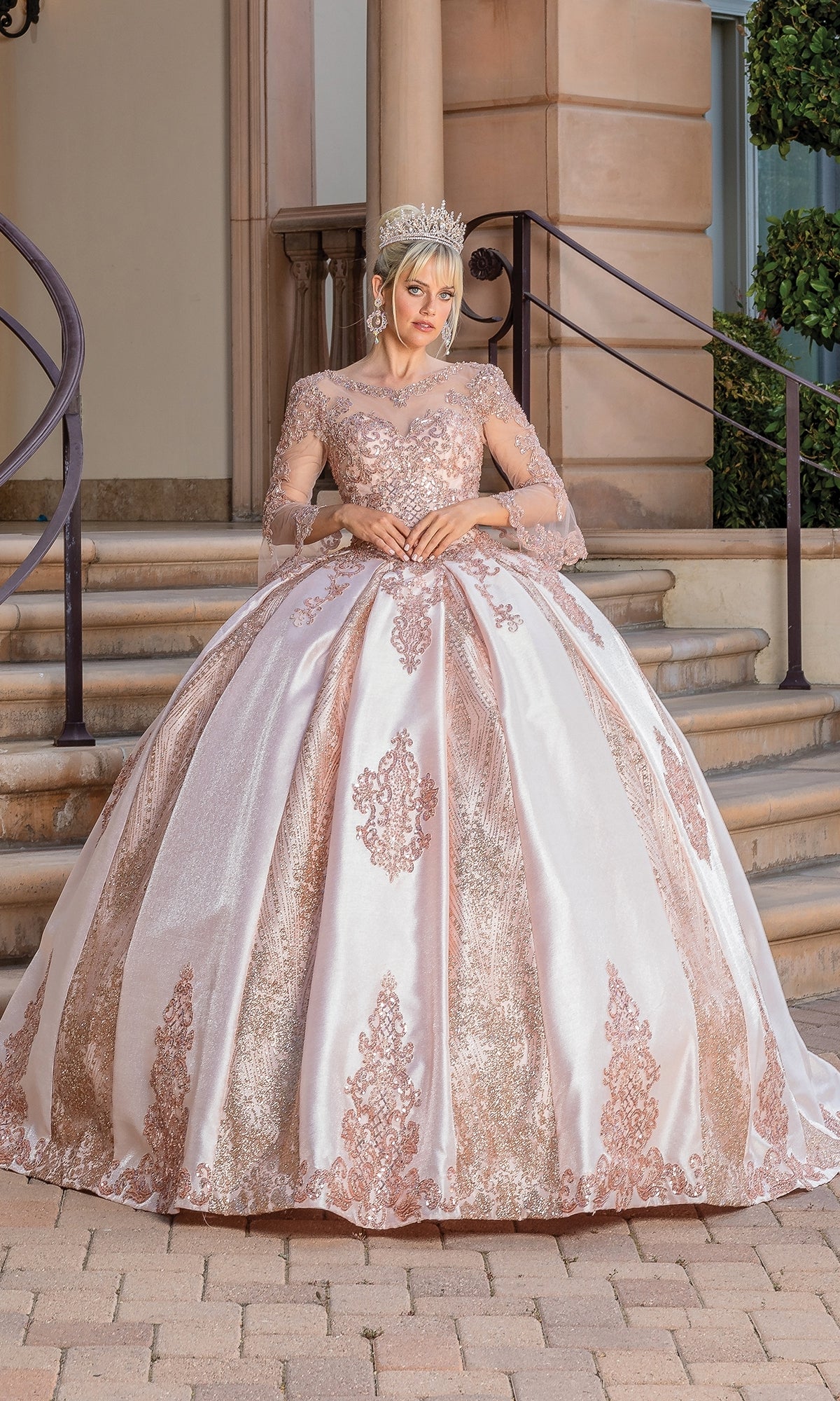 A woman poses on stone steps before a grand building, wearing Quinceanera Dress 1665 By Dancing Queen—a sparkling ball gown with sheer bell sleeves and a sweetheart neckline.