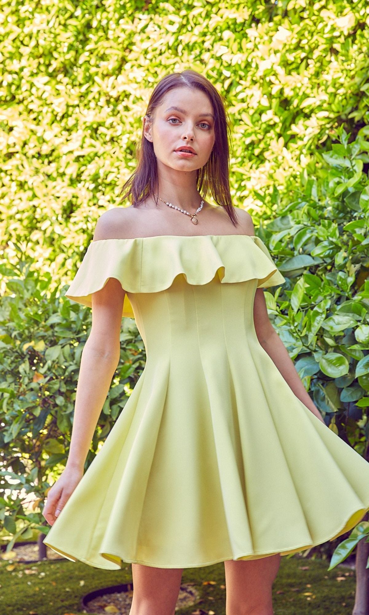 A woman poses outdoors against lush green foliage, wearing the affordable Off-the-Shoulder Short Party Dress in light yellow, featuring ruffles and a flared skirt.