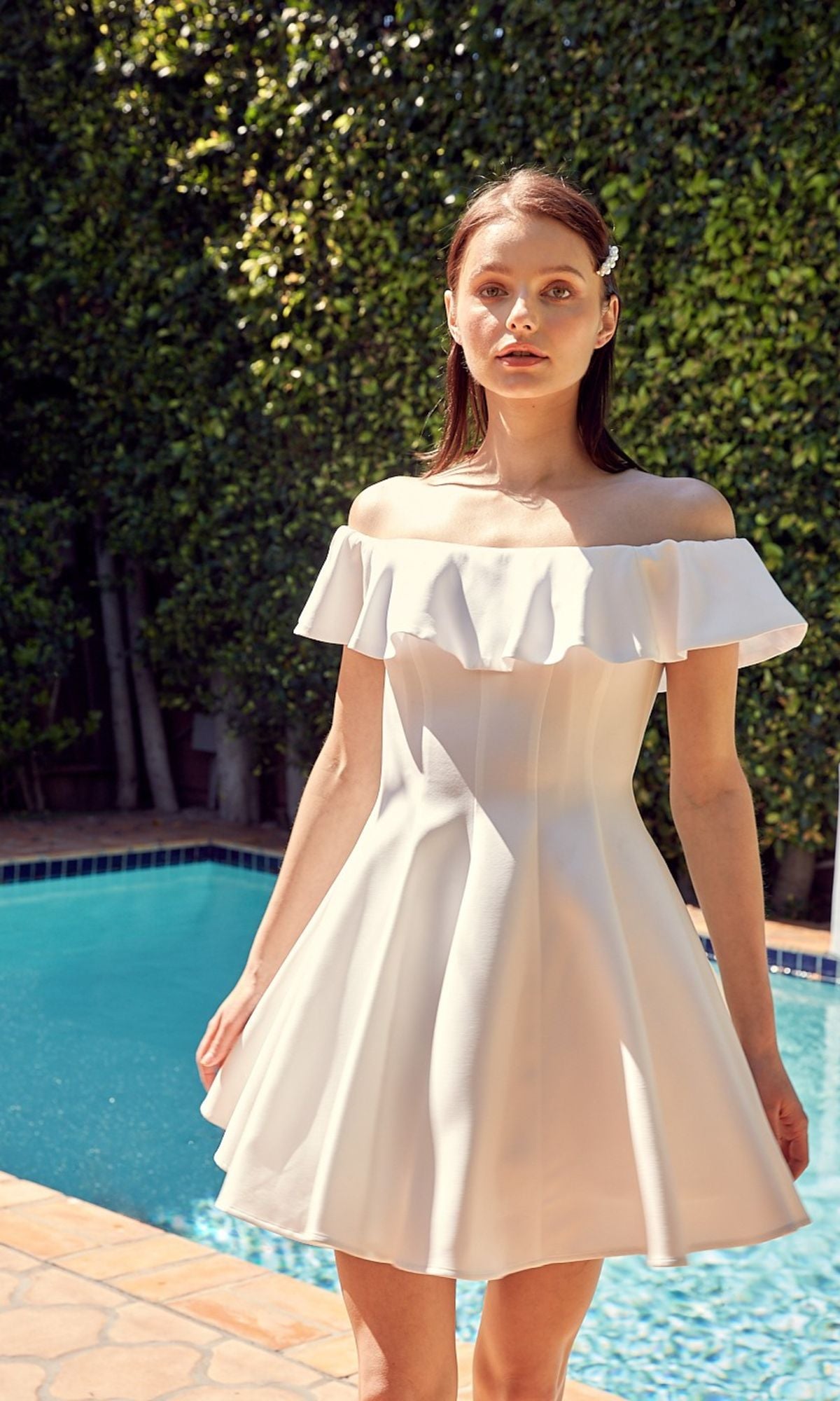 A woman in an Off-the-Shoulder Short Party Dress poses by a swimming pool, with vibrant green foliage providing a lush backdrop.