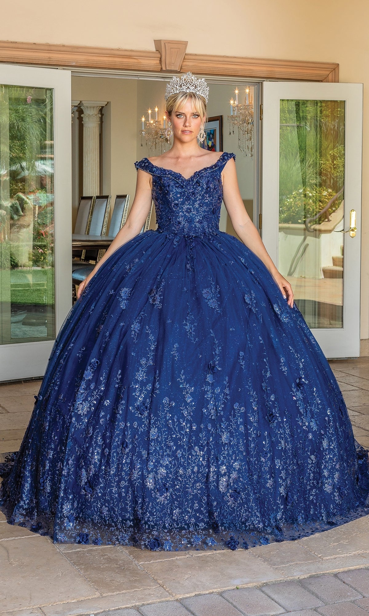 A woman wearing Quinceanera Dress 1668 by Dancing Queen—a blue ball gown with a sweetheart neckline, full skirt, and floral designs—stands indoors in front of open glass doors, crowned elegantly.