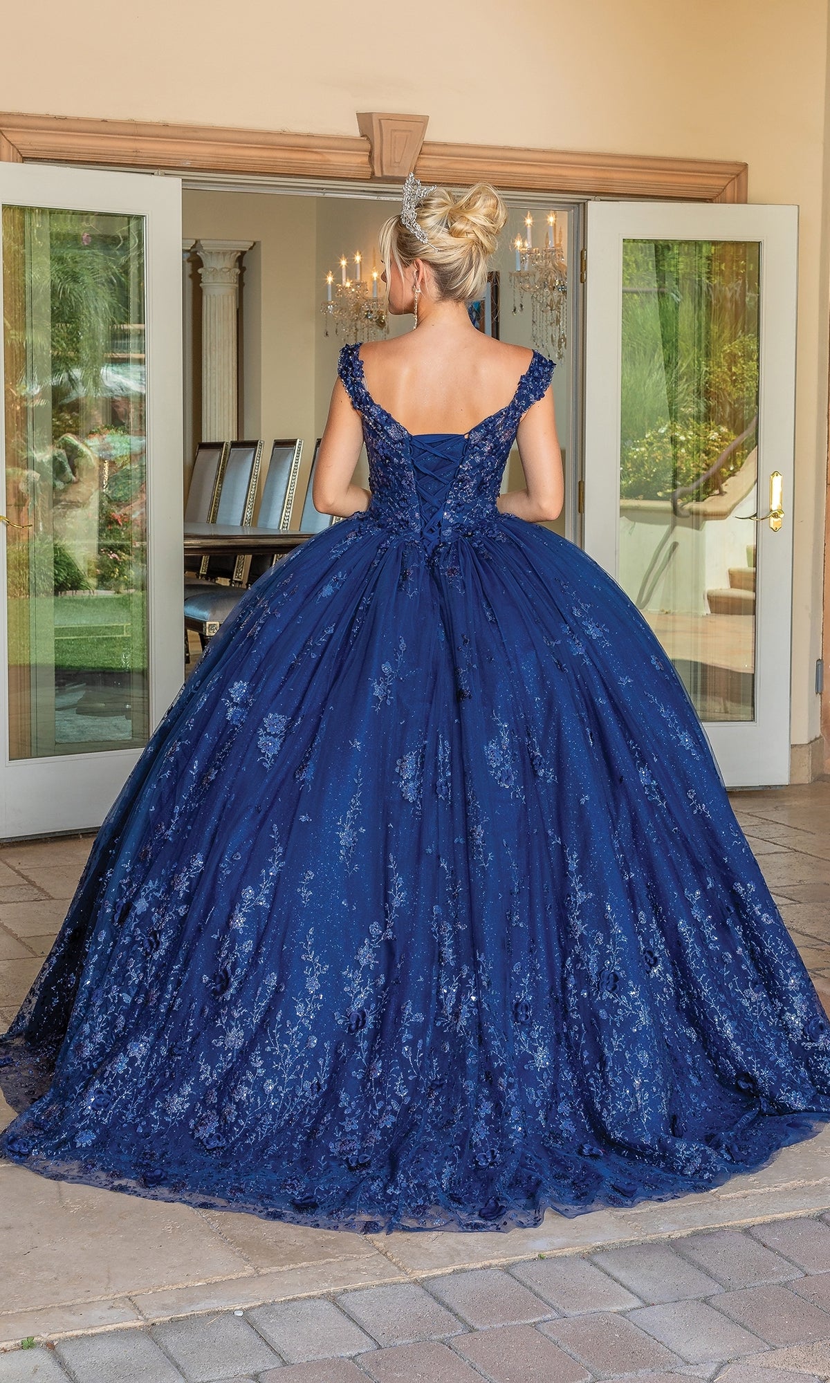 A woman faces away from the camera in the Quinceanera Dress 1668 By Dancing Queen, a navy blue, lace-detailed A-line ball gown with a sweetheart neckline, standing outside an open doorway.