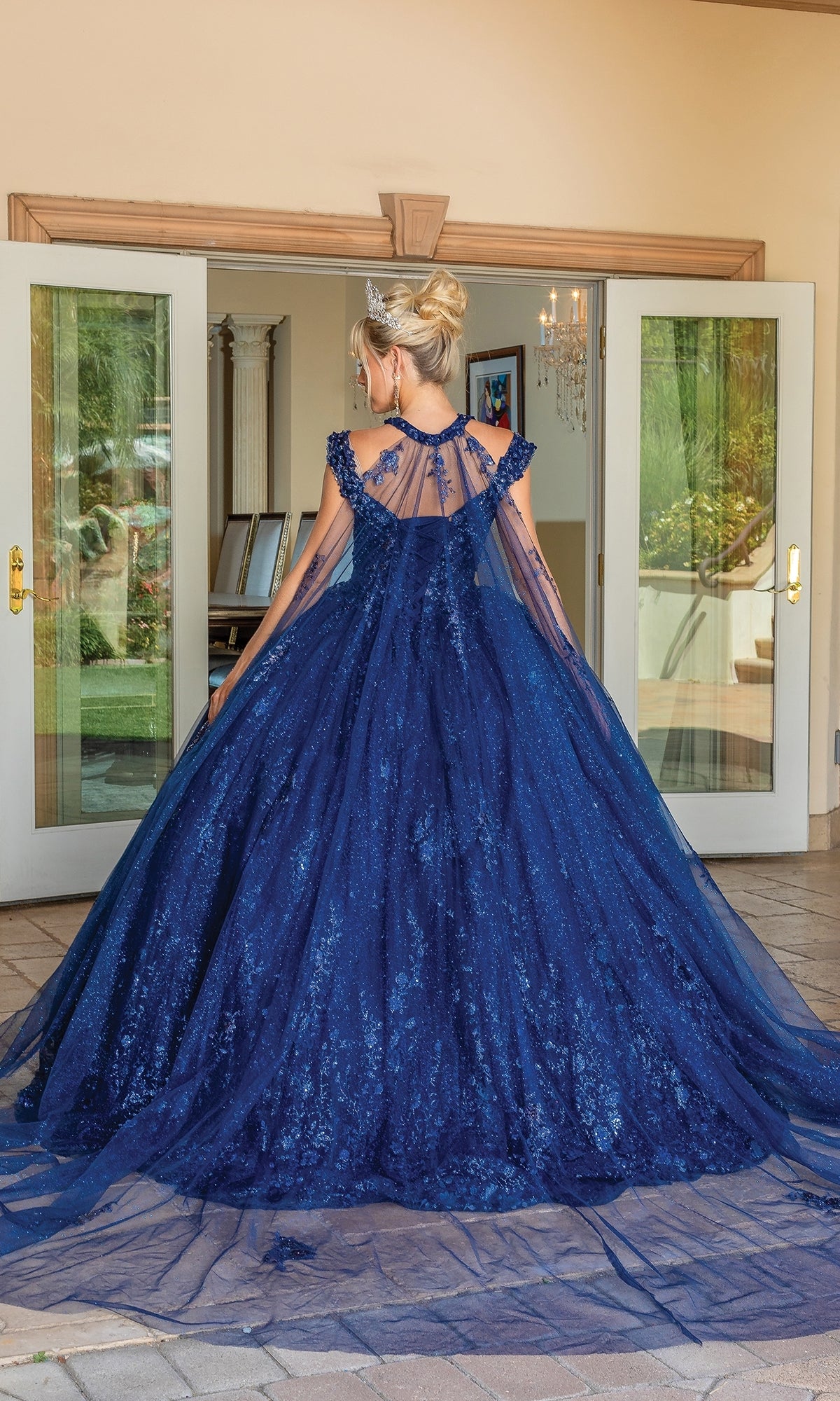 A woman stands outdoors facing away, wearing the Quinceanera Dress 1668 By Dancing Queen—a floor-length, dark blue, glittering ball gown with a sheer cape, sweetheart neckline, and open back. French doors and greenery appear in the background.