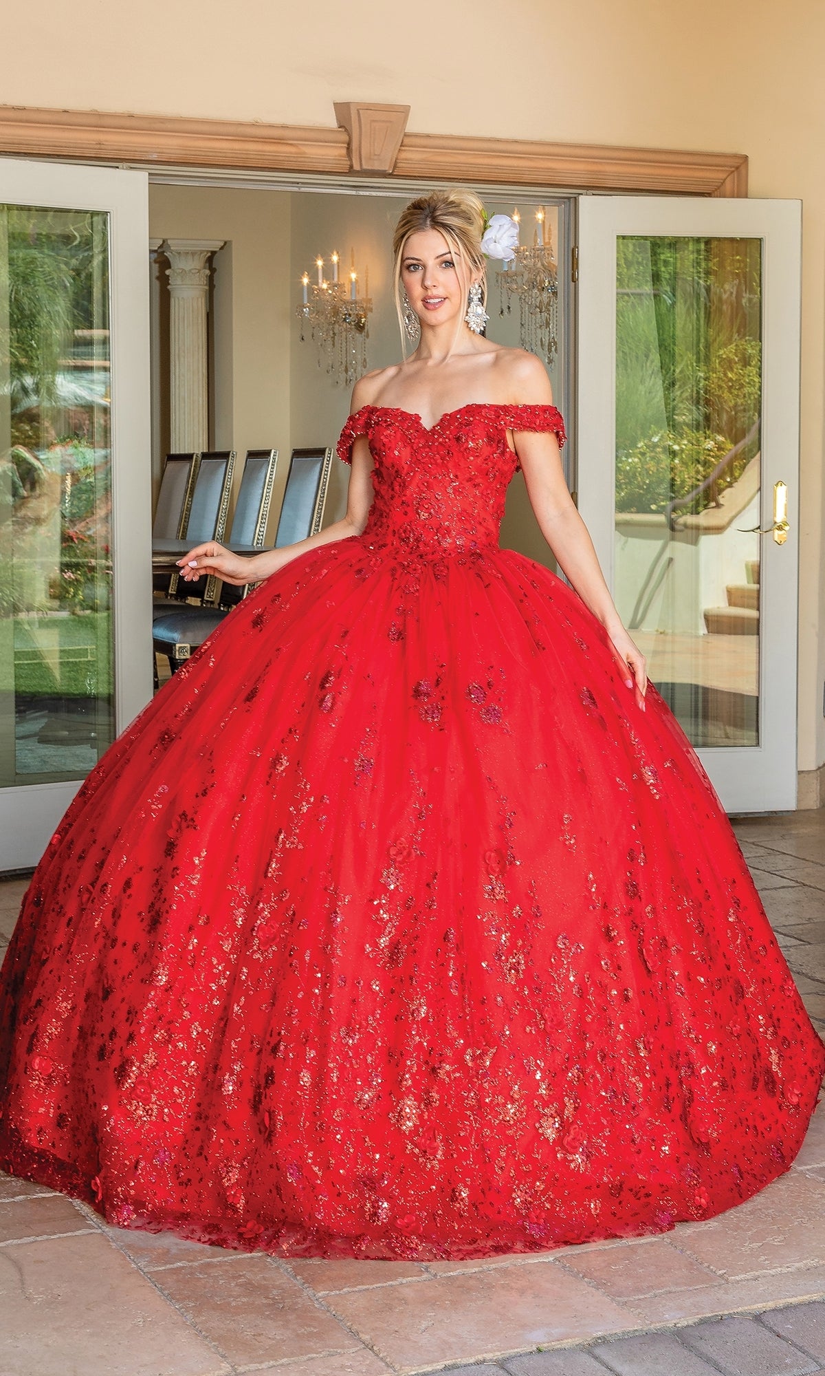 A woman wearing the Quinceanera Dress 1668 By Dancing Queen, an off-the-shoulder A-line ball gown with floral embellishments, stands before open glass doors, chandelier and greenery visible inside.