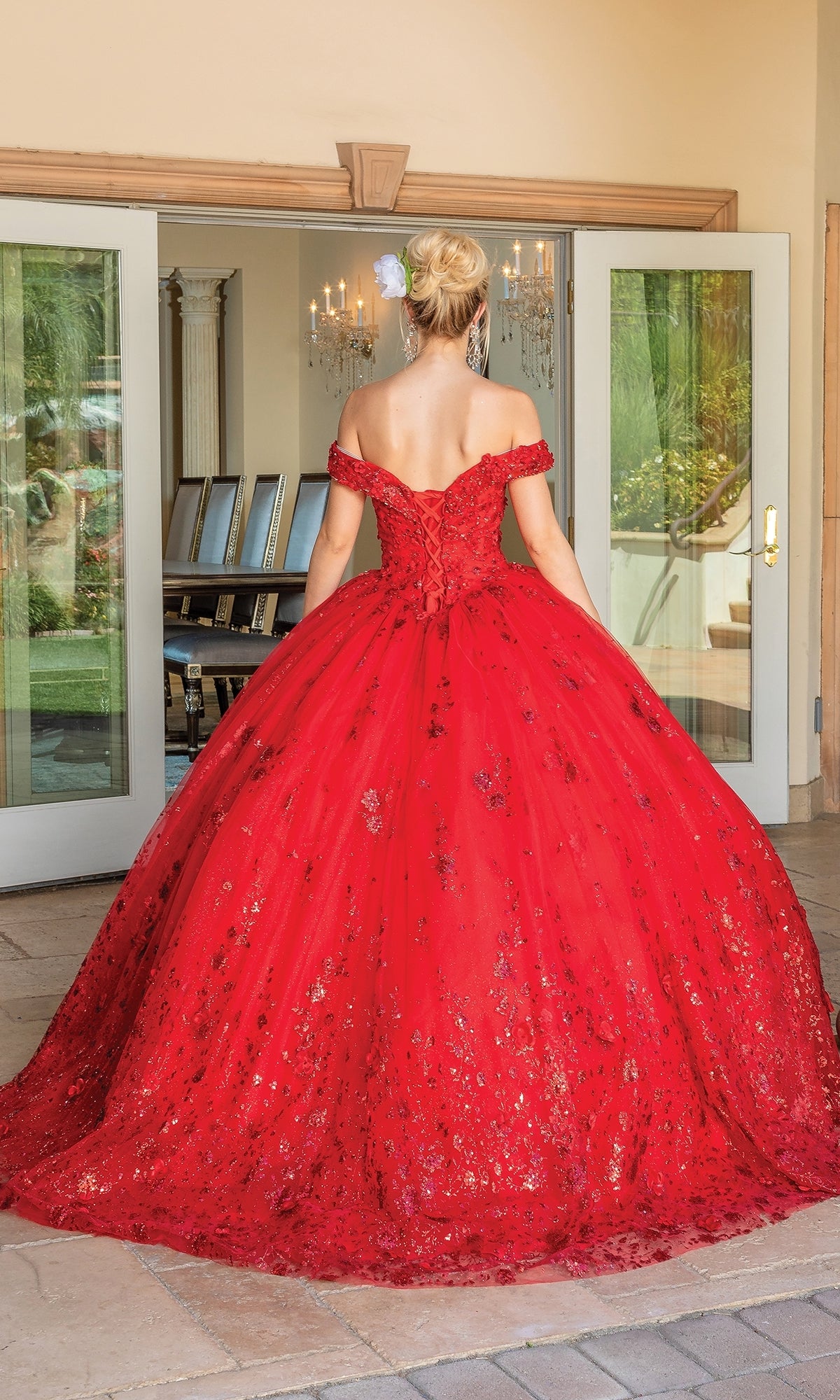 A woman in the Quinceanera Dress 1668 by Dancing Queen, a floor-length red ball gown with floral details and an off-the-shoulder sweetheart neckline, stands facing away in front of glass doors.