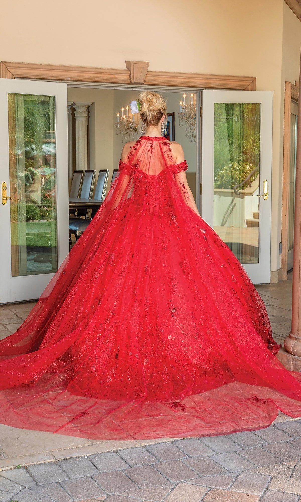 A person stands outside double doors in the Quinceanera Dress 1668 By Dancing Queen—a floor-length, bright red ball gown with a sweetheart neckline and long, sheer embellished train—at an elegant venue.