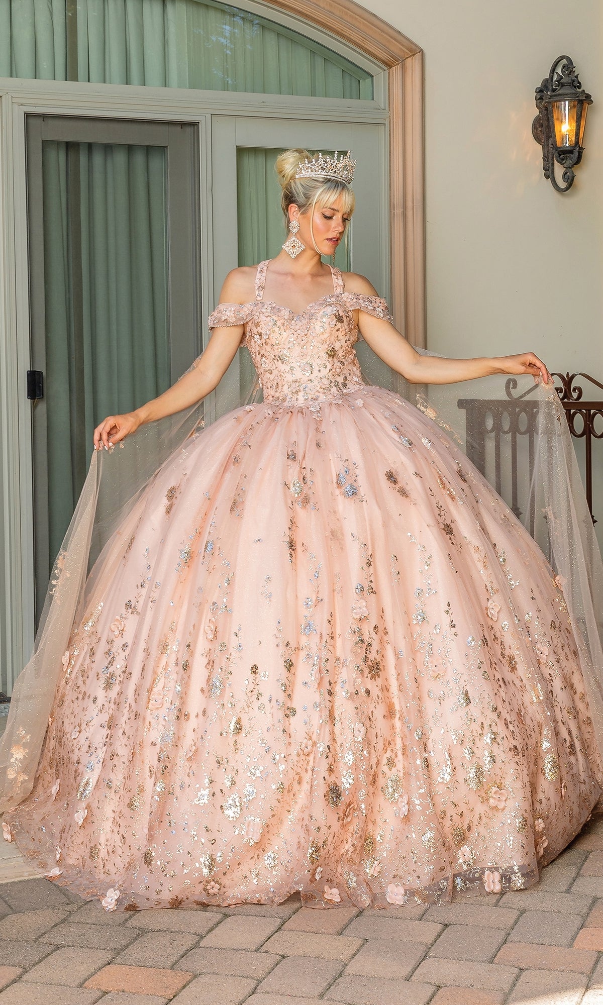 Wearing the Quinceanera Dress 1668 by Dancing Queen, a person in a pink, embellished ball gown with off-shoulder sleeves and sweetheart neckline stands outside in front of large windows and a light fixture, accessorized with a tiara.