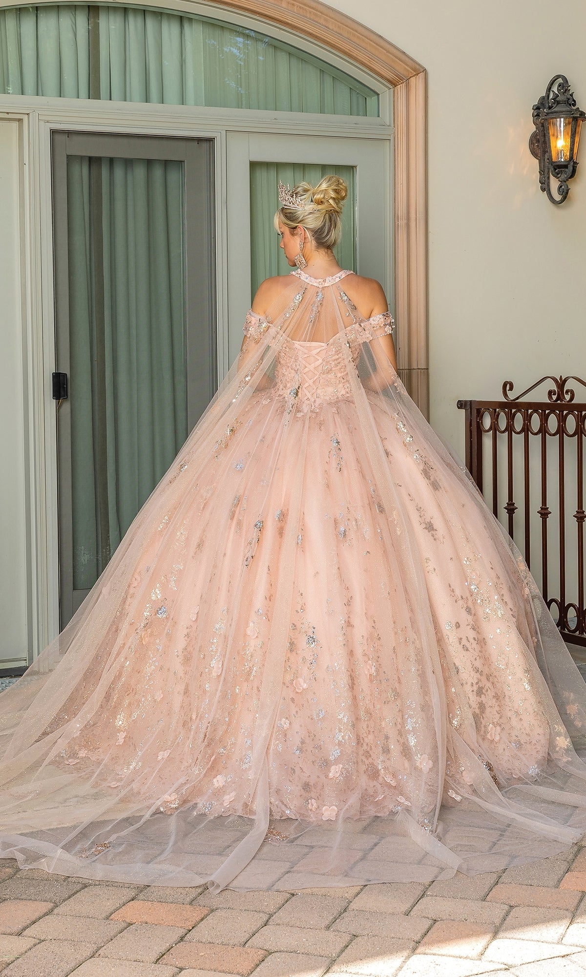 A woman in the Quinceanera Dress 1668 By Dancing Queen, a light pink embellished ball gown with a cape and tiara, stands facing away in front of glass doors.