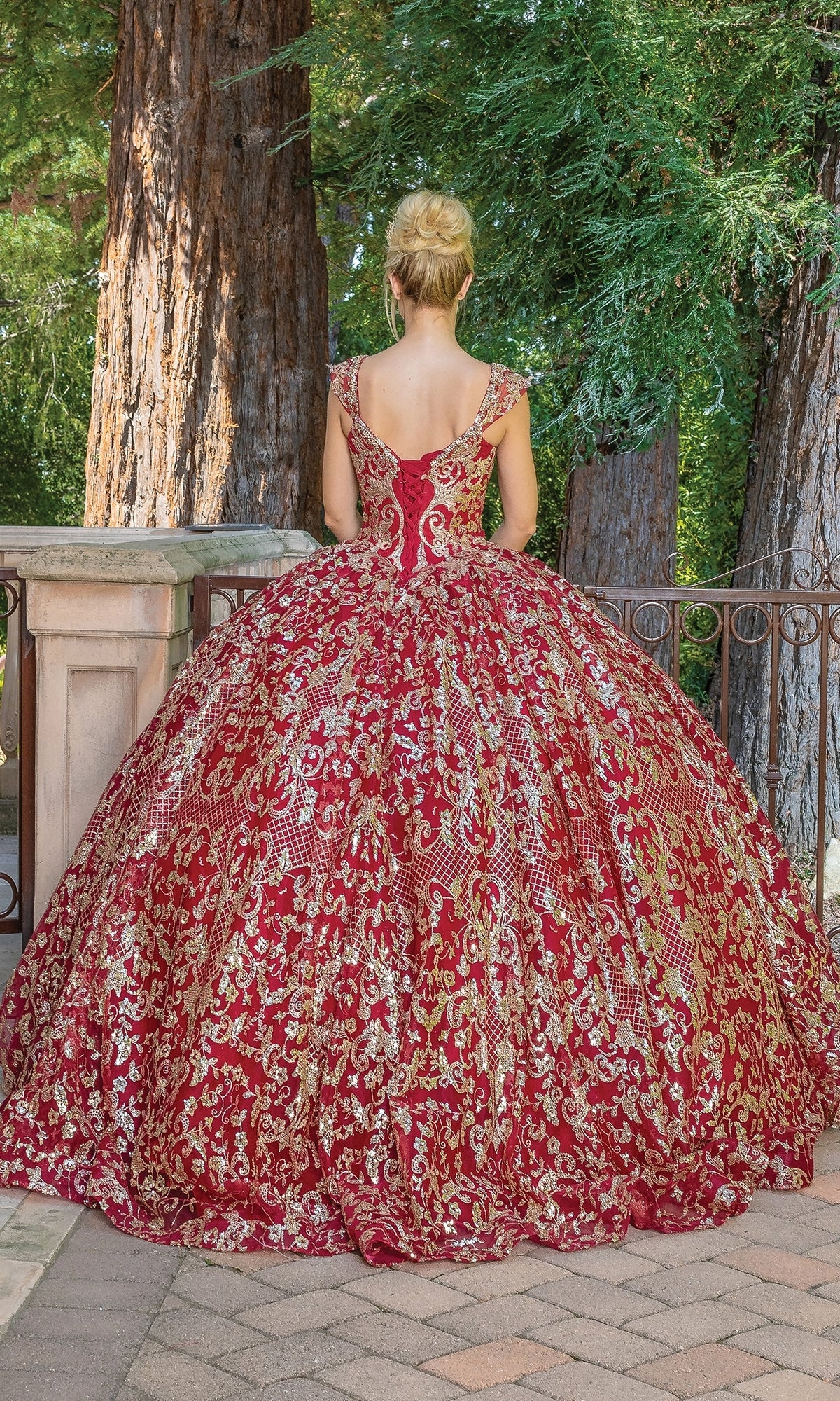 A woman wearing the Quinceanera Dress 1670 By Dancing Queen stands outdoors with her back to the camera, facing a railing and towering trees.