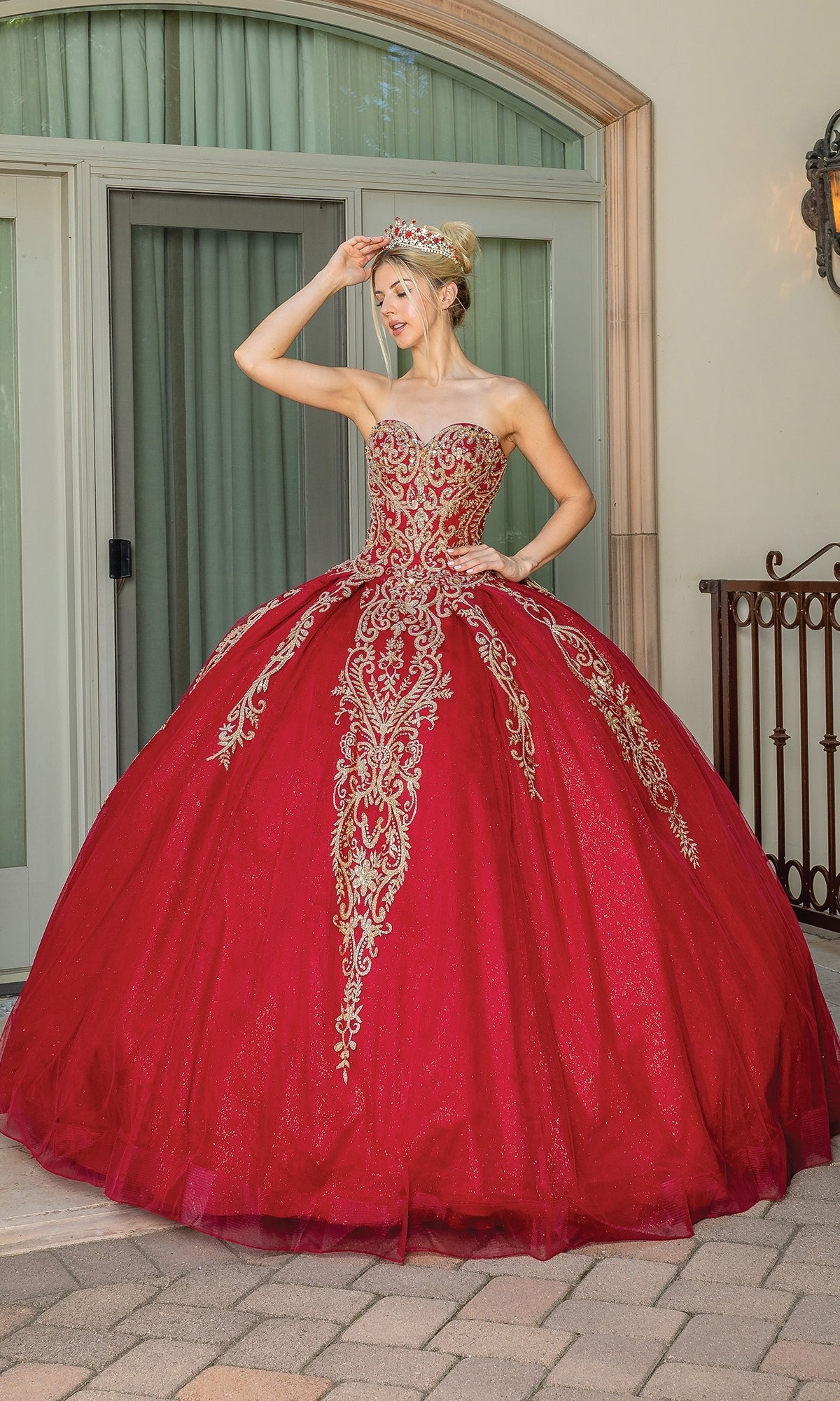 Model wears the Traditional Quinceañera Dress: Dancing Queen 1672—a strapless red ball gown with gold embroidery—outside, posing with one hand on her head and accessorized with a jeweled crown.