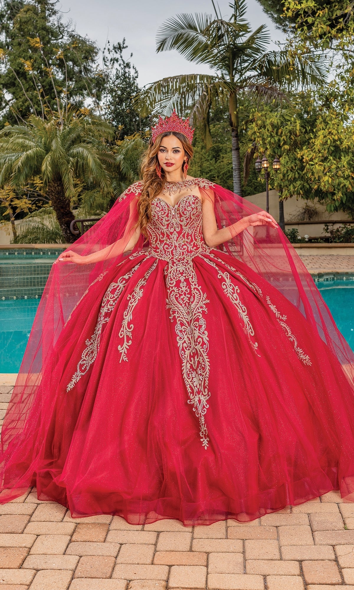 A woman stands by a pool in the Dancing Queen 1672 Traditional Quinceañera Dress, a red ball gown with silver embroidery, matching cape, and jeweled crown. Palm trees decorate the background.