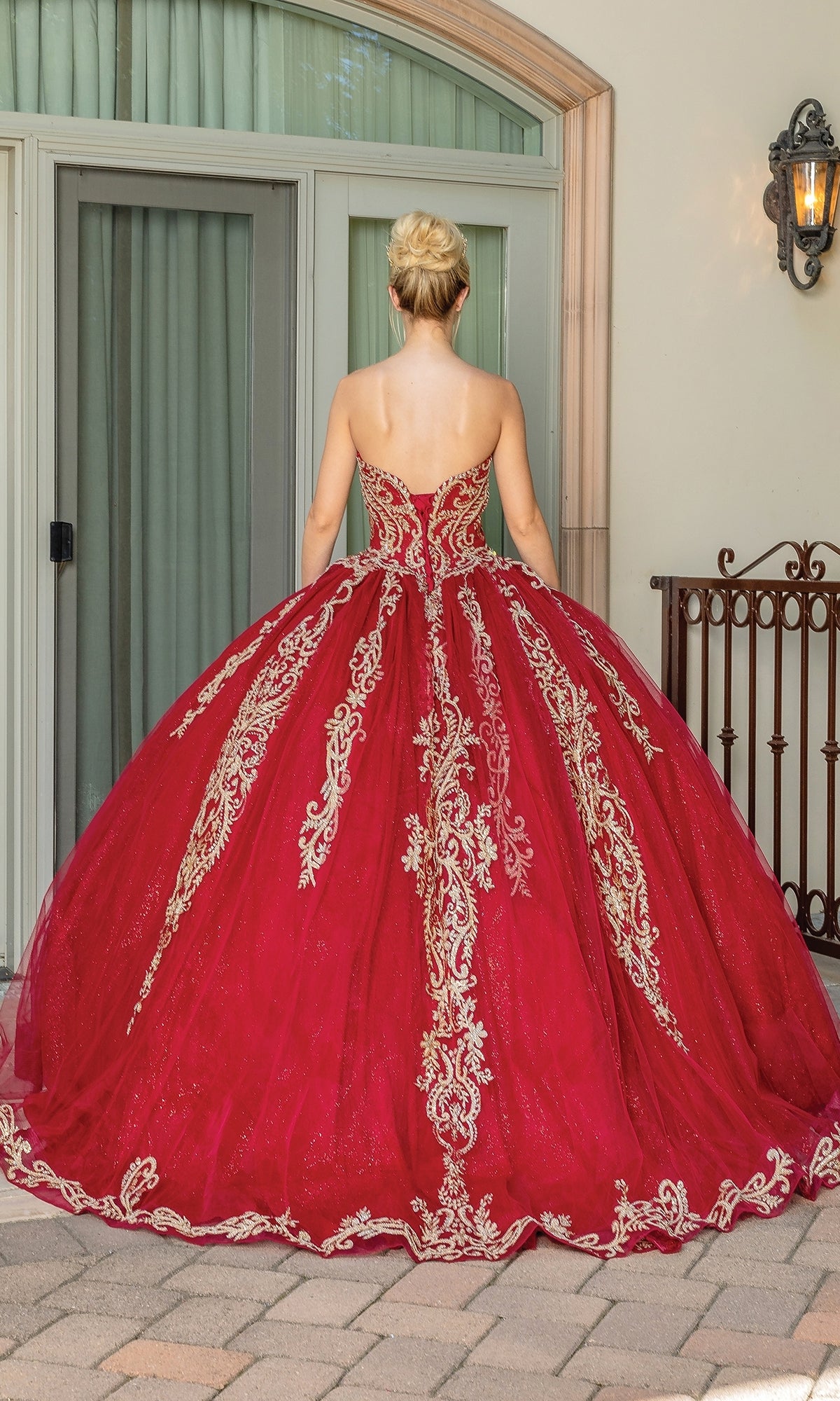 A woman wears the Traditional Quinceañera Dress: Dancing Queen 1672—a red ball gown with gold embroidery—standing on a paved patio in front of a glass door, facing away.