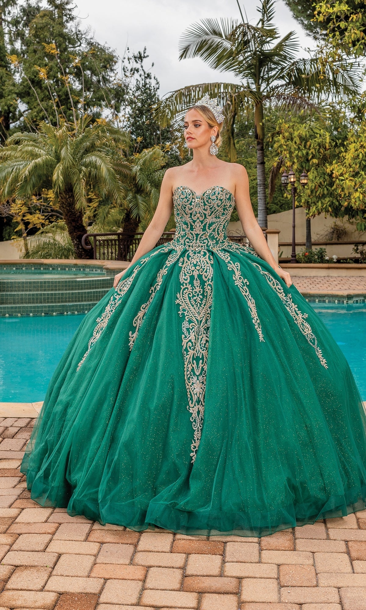 A woman wearing the Dancing Queen 1672 traditional quinceañera dress poses by a pool lined with palm trees, stunning in a green strapless ball gown accented with silver embroidery.