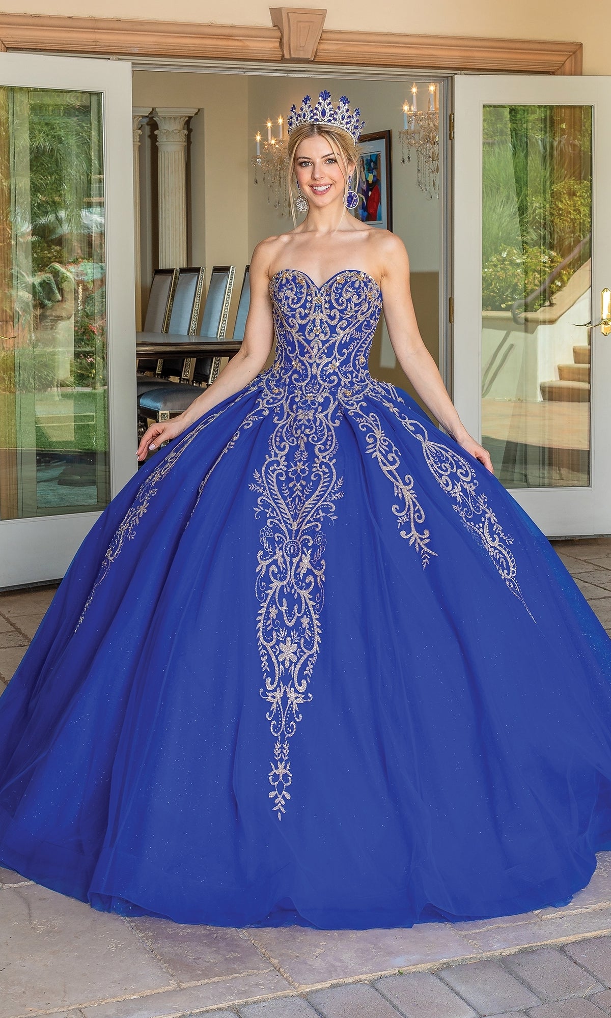 A woman in the Traditional Quinceañera Dress: Dancing Queen 1672, a royal blue ball gown with intricate silver embroidery and a jeweled crown, stands smiling in front of glass doors.