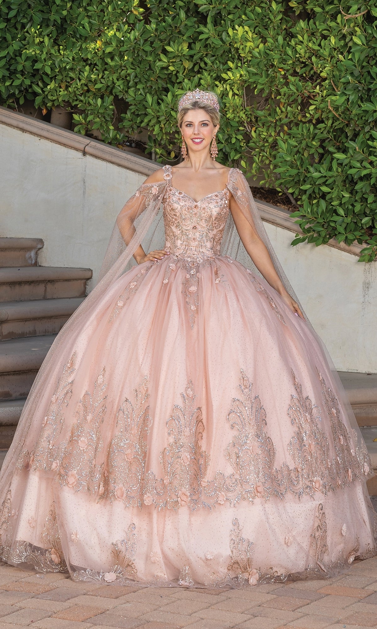 A woman in the Dancing Queen 1676 Quince Dress with Cape Sleeves, featuring ornate blush pink embroidery and a tiara, stands on a paved path before green foliage and stairs.