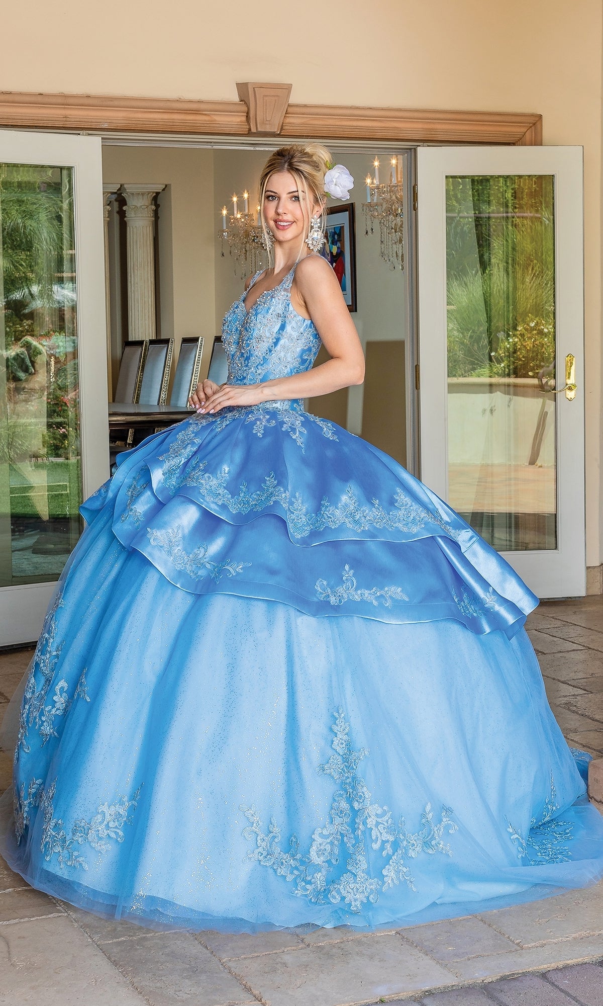 A young woman stands outside before French doors, wearing the Quinceanera Dress 1682 by Dancing Queen, featuring lace detailing, a sweetheart neckline, and a flower headpiece.