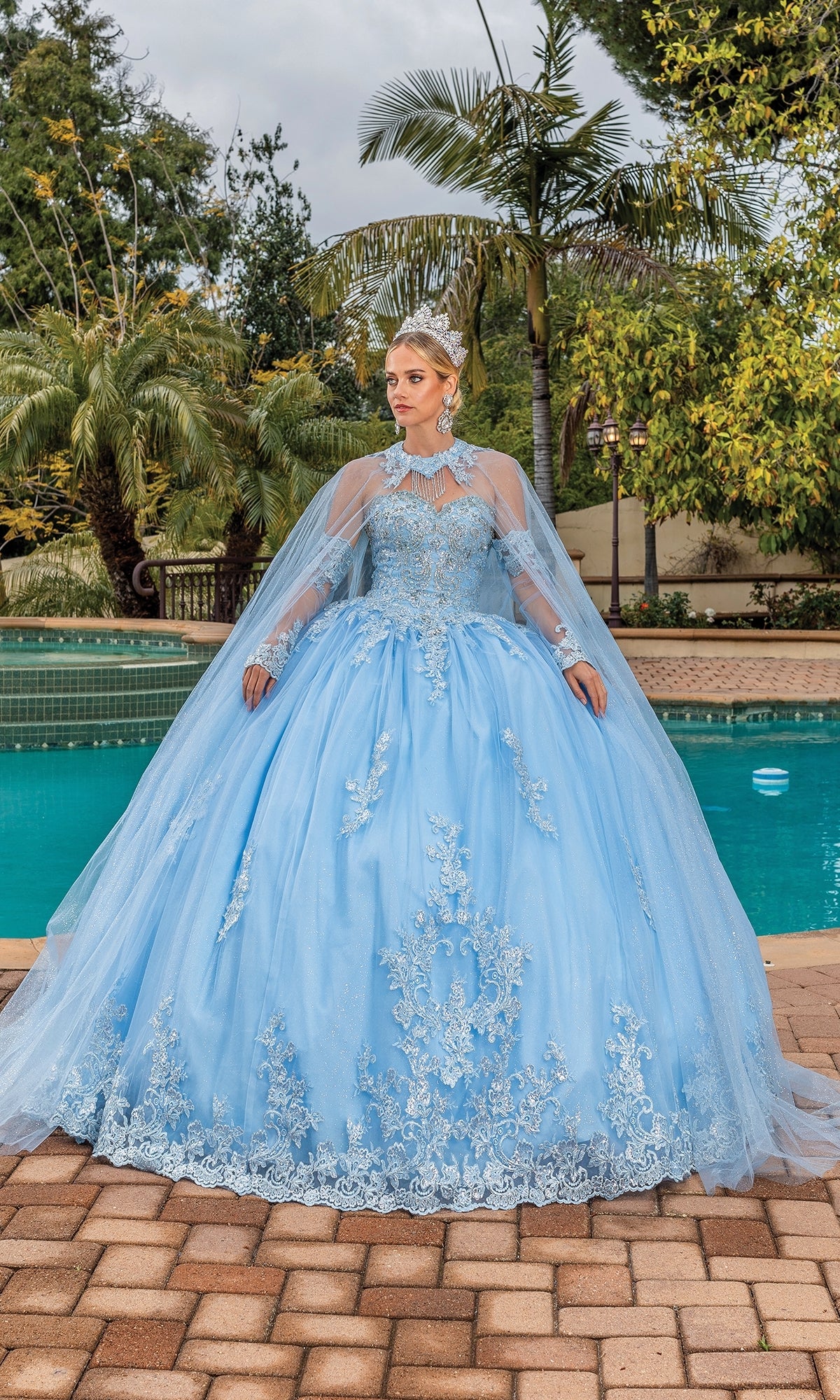 A woman stands by a pool wearing the Quinceanera Dress 1691 by Dancing Queen, a light blue strapless ball gown with lace details, removable cape and sleeves, elegantly accented with a tiara.