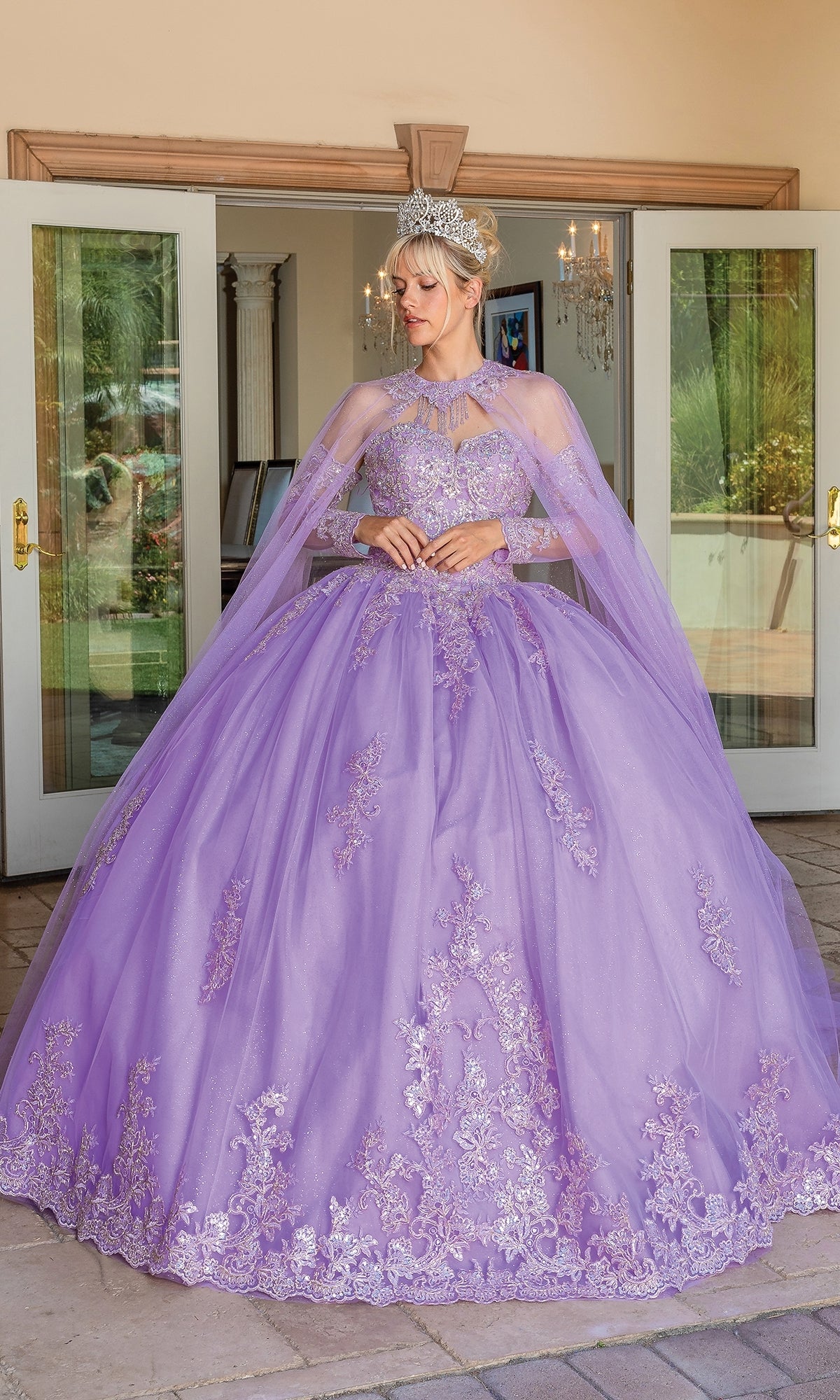 A woman in the Quinceanera Dress 1691 By Dancing Queen, a strapless lilac ball gown with lace details and a removable cape, stands before open glass doors, wearing a tiara.
