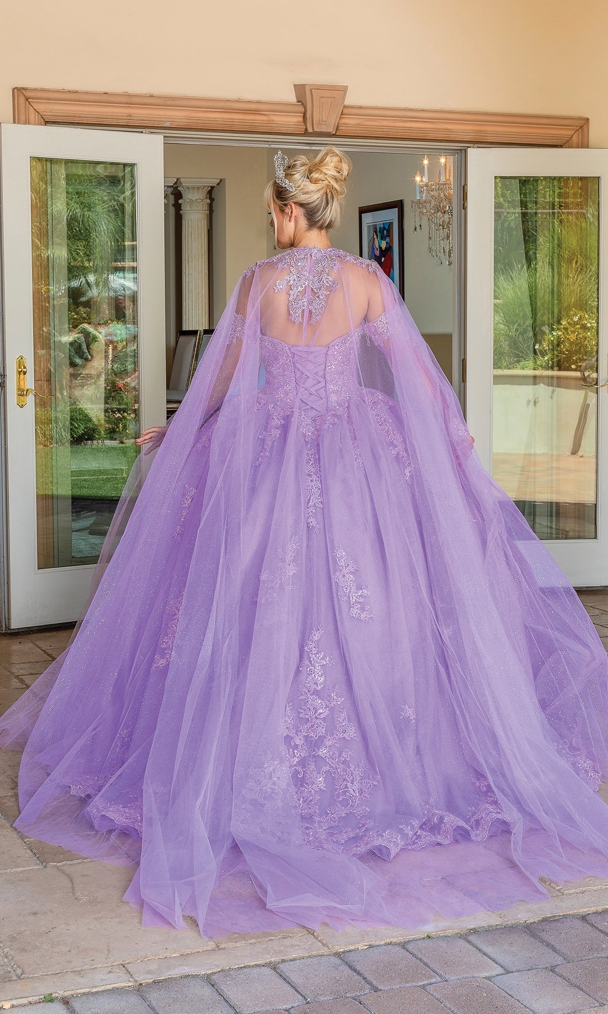 A person stands facing away in front of open double doors, wearing Quinceanera Dress 1691 By Dancing Queen—a floor-length, light purple strapless ball gown with lace details and a sheer removable cape.