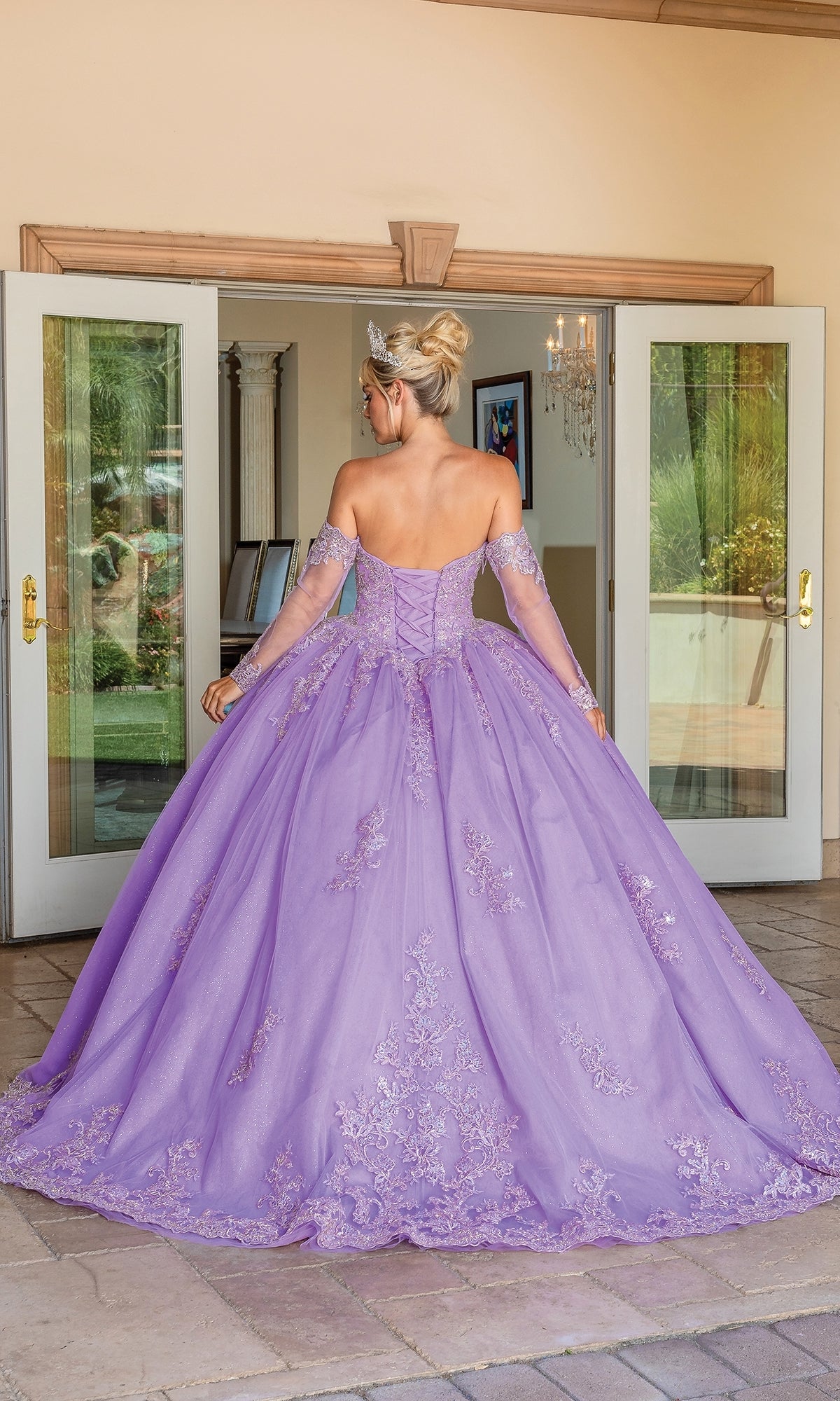 A woman wearing the Quinceanera Dress 1691 By Dancing Queen, a lavender off-the-shoulder embroidered A-line gown, stands facing away in front of open double doors with a tiara and her hair in an updo.