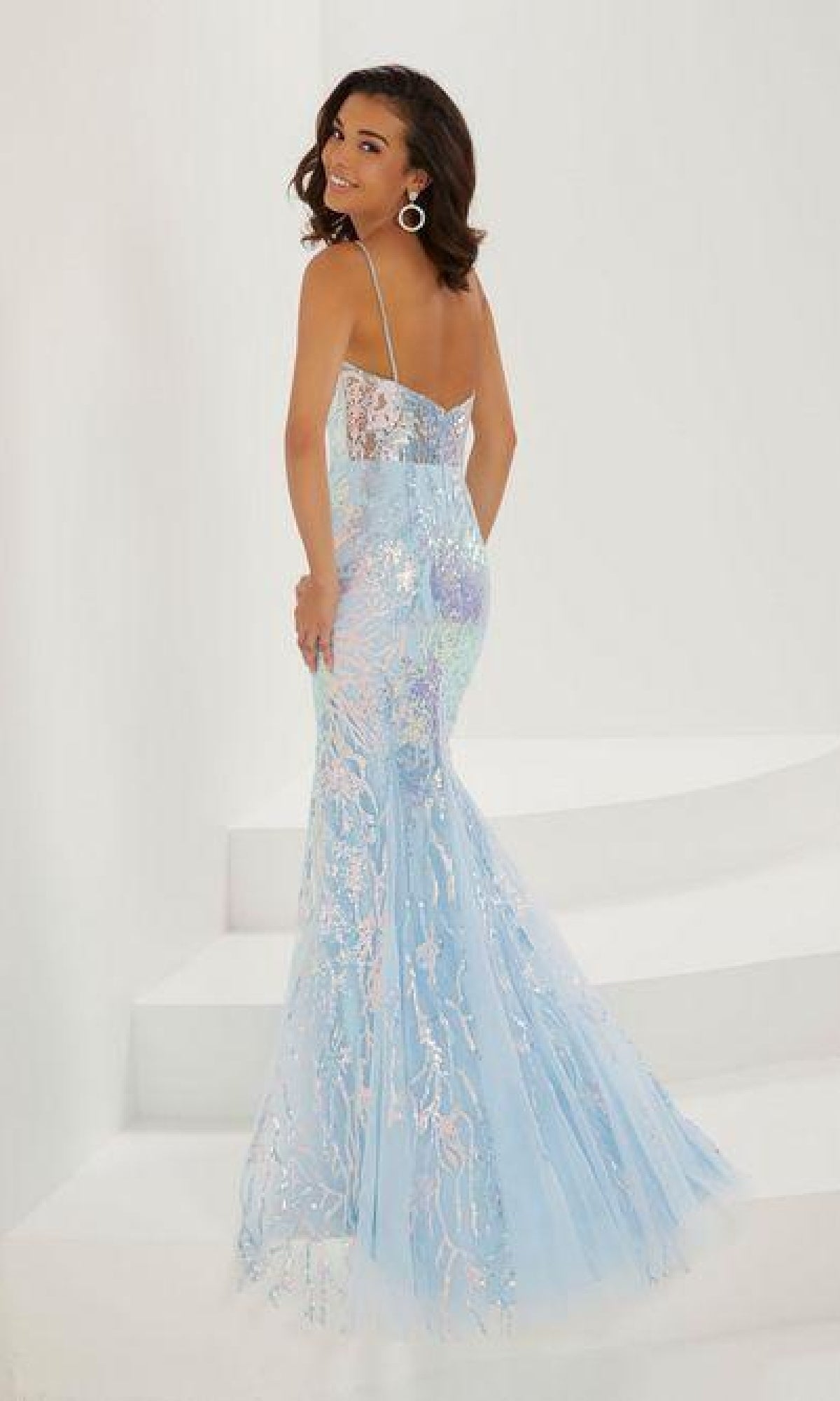 Wearing the Long Sequin Prom Dress: Tiffany 16944, a woman with thin straps and a tulle overlay stands on white steps, glancing back over her shoulder.