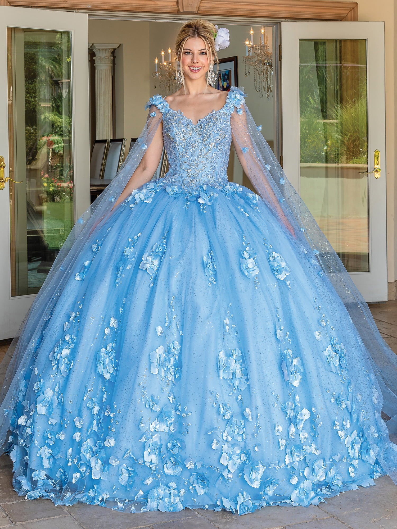 A woman in the Quinceanera Dress 1695 By Dancing Queen, featuring a beautiful Bahama Blue color and elegant cape sleeves, stands smiling in a doorway. Surrounded by chandeliers and lush greenery, delicate floral details accentuate her enchanting a-line ball gown look.