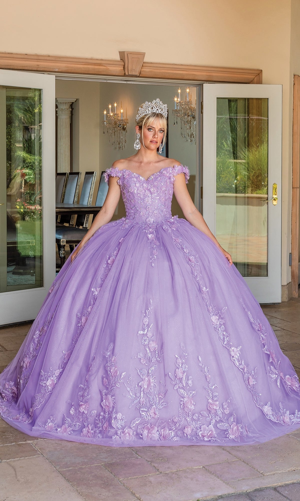 A person wearing the Quinceanera Dress 1698 By Dancing Queen, a detailed floral ball gown with a sweetheart neckline, and a tiara stands in front of open glass doors.