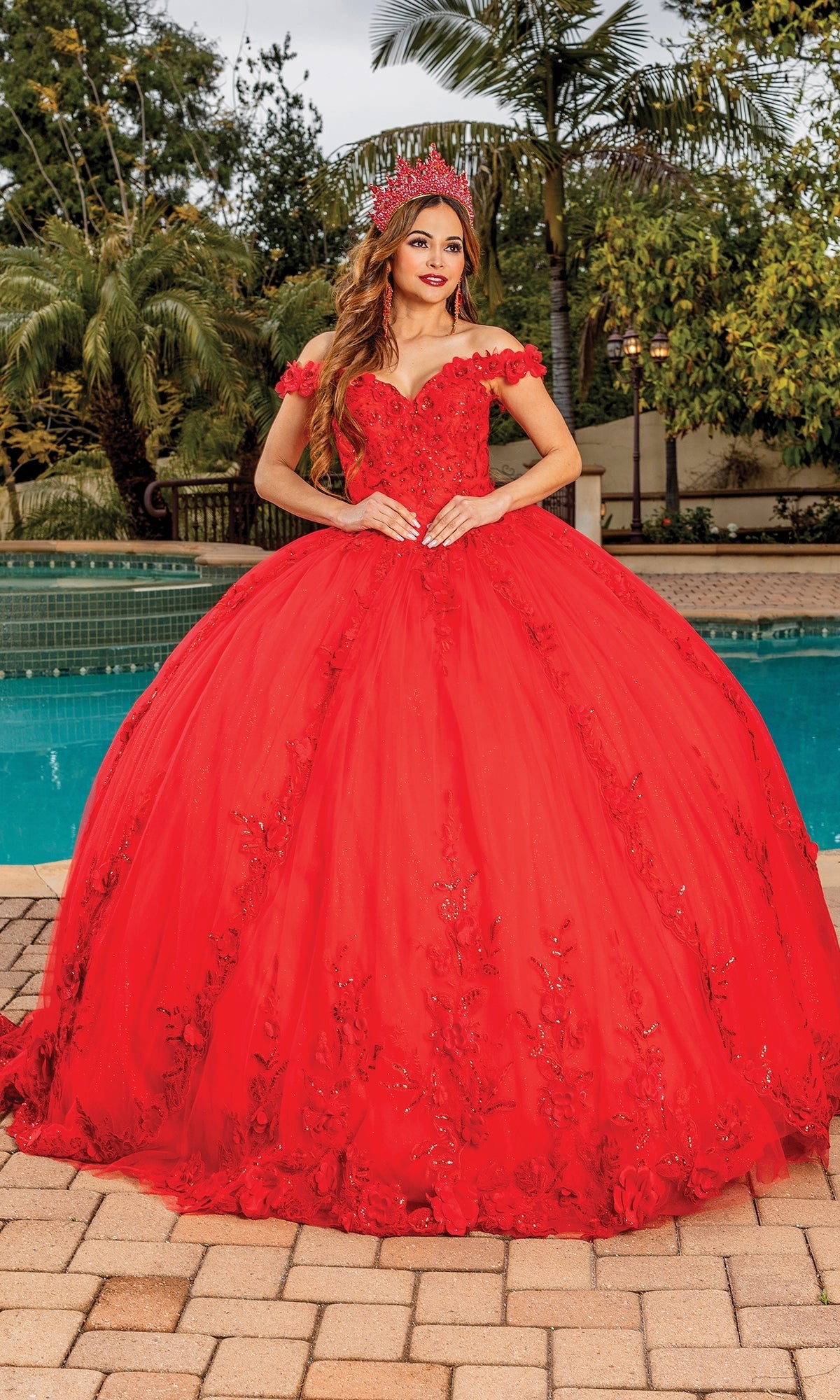 A woman stands outdoors by a pool, wearing the Quinceanera Dress 1698 By Dancing Queen, a bright red off-the-shoulder gown with floral detailing and a matching red crown.