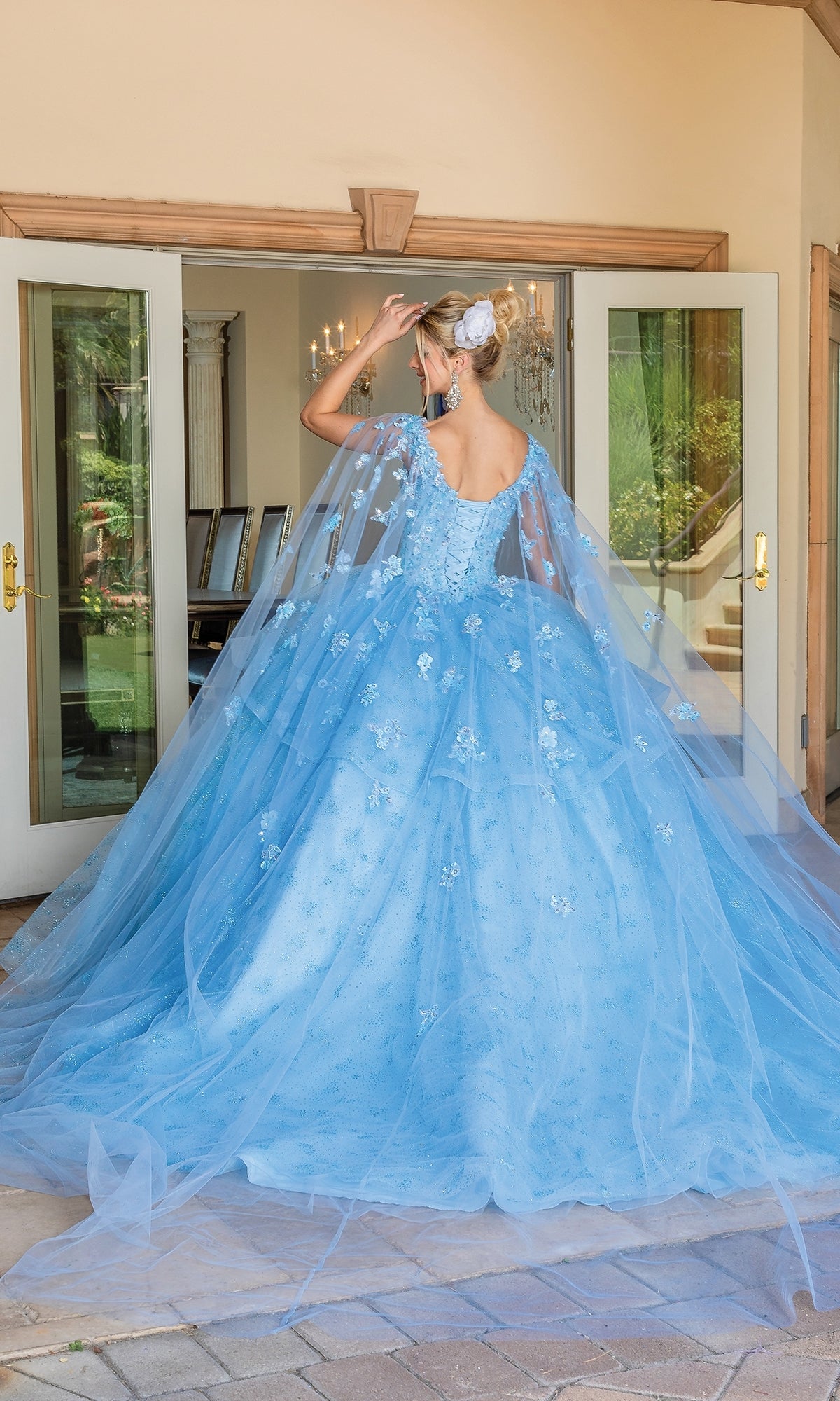 A woman in the Quinceanera Dress 1702 By Dancing Queen—a light blue, floral-embroidered A-line ball gown with a v-neck and long tulle train—stands facing away before open double doors.