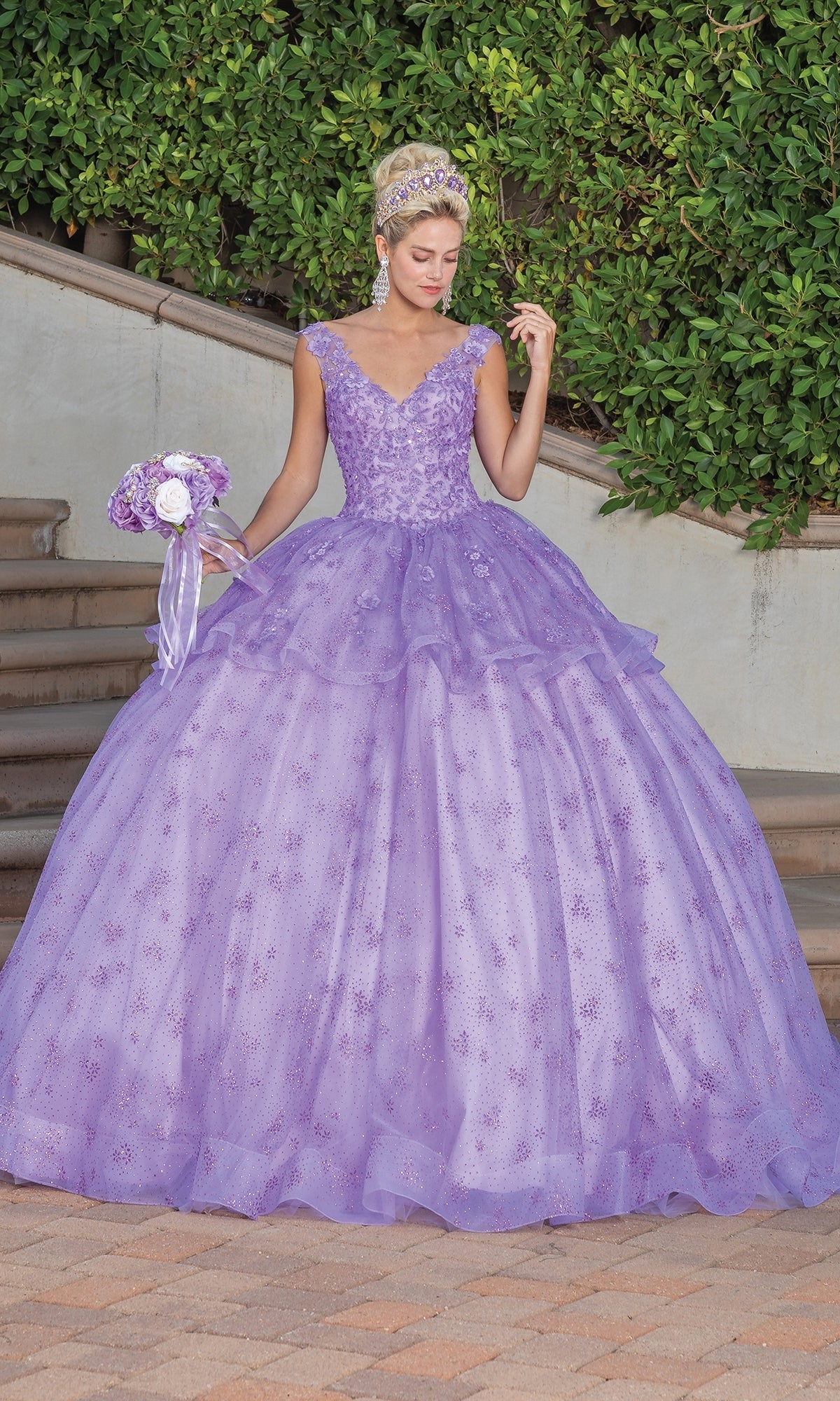 A woman wearing the Quinceanera Dress 1702 By Dancing Queen—a lavender ball gown with a full skirt and lace embellished cape—stands outdoors holding a matching bouquet, with greenery and steps in the background.