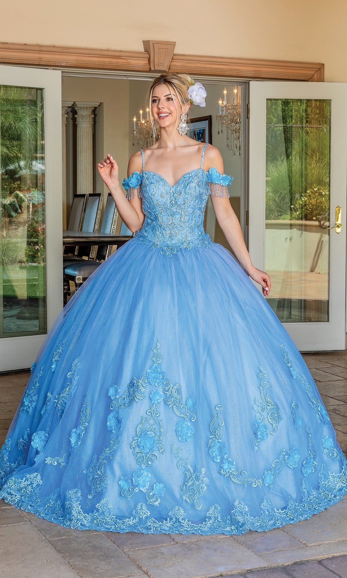 A woman stands indoors wearing the Quinceanera Dress 1706 By Dancing Queen, a blue off-the-shoulder ball gown with lace details and a full skirt, smiling with one hand raised.