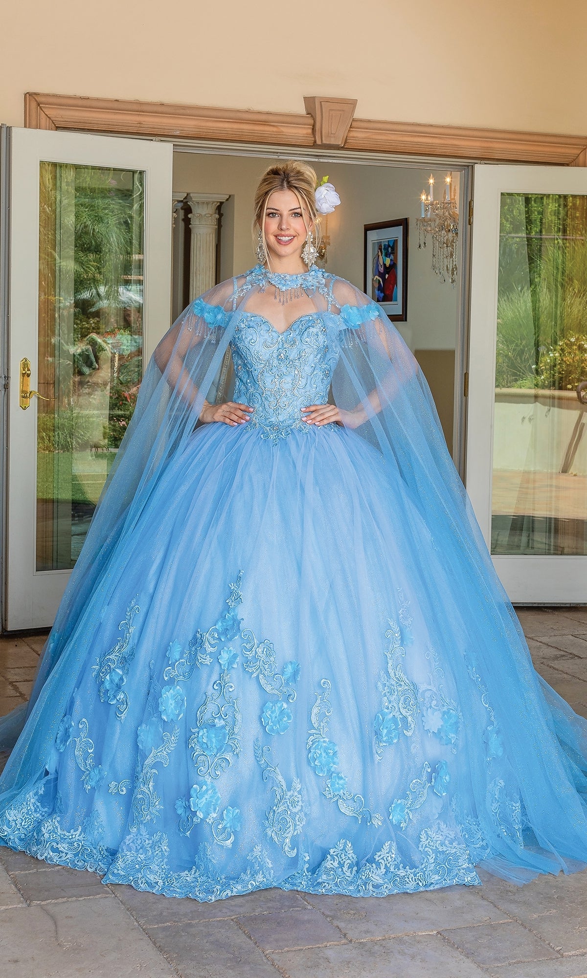 A woman stands outside double doors wearing the Quinceanera Dress 1706 By Dancing Queen, featuring off-the-shoulder floral embroidery, sheer removable cape sleeves, and a matching floral hair accessory.
