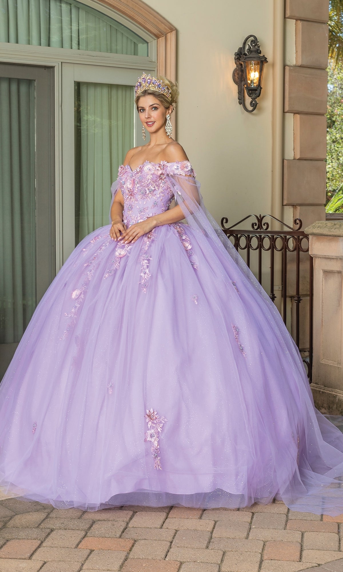 A woman wears the Quinceanera Dress 1714 By Dancing Queen, a lilac A-line gown with floral details and a crown, standing outside a building with large windows and stone columns.