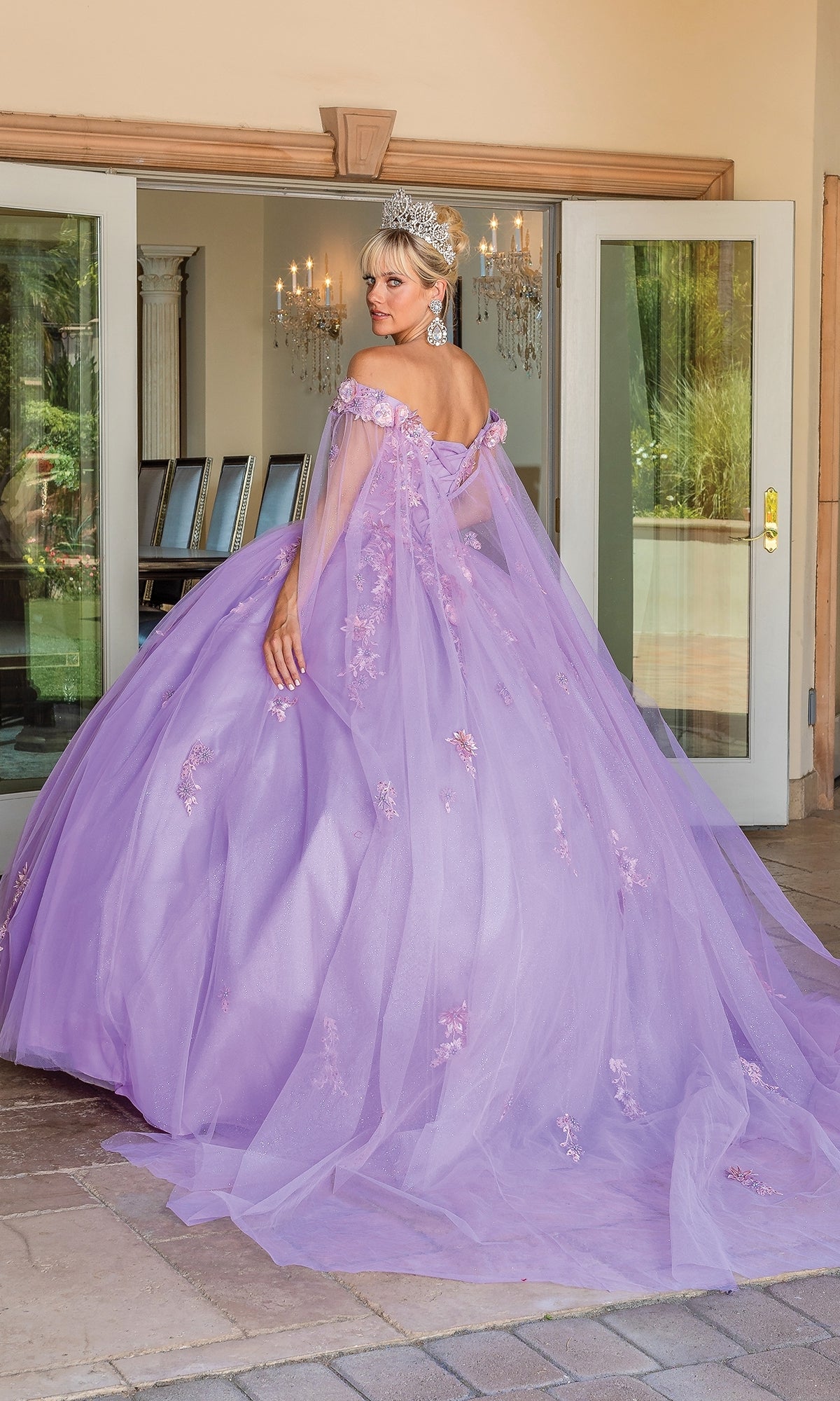 A woman in the Quinceanera Dress 1714 By Dancing Queen and a tiara stands outdoors, facing away, with glass doors and chandeliers visible in the background.
