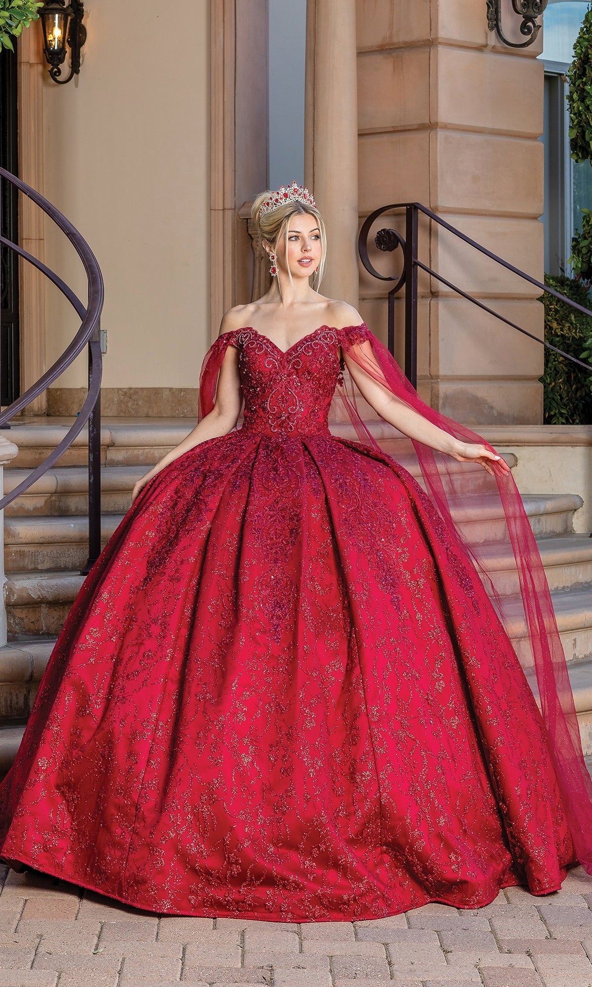 A woman wearing the Burgundy Red Quinceanera Dress 1722, featuring an off-the-shoulder design and crown, stands on a stone walkway before steps and a building entrance.