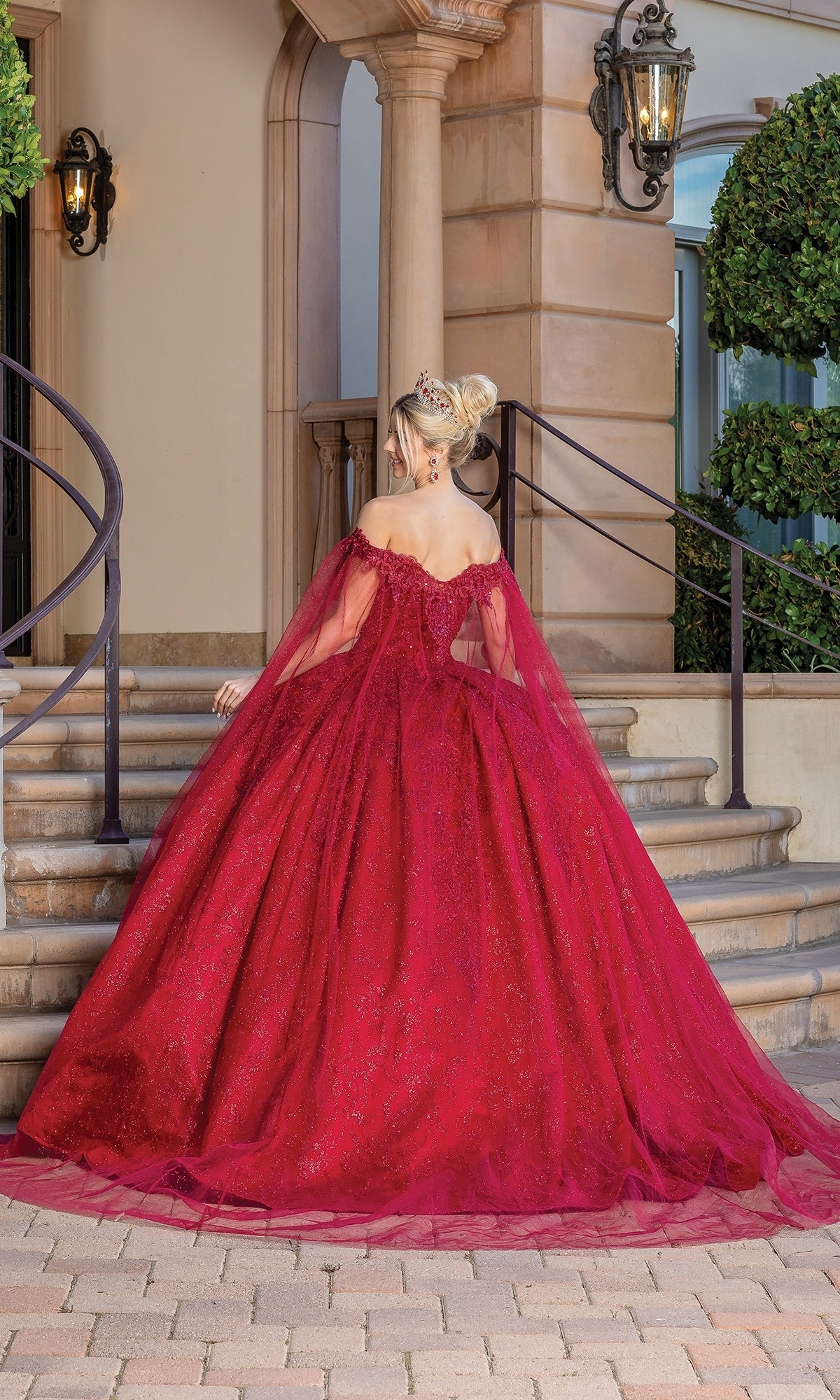 Wearing the Burgundy Red Quinceanera Dress 1722, a woman stands on stone steps facing away, showcasing the gown’s voluminous off-the-shoulder style and dramatic long train.