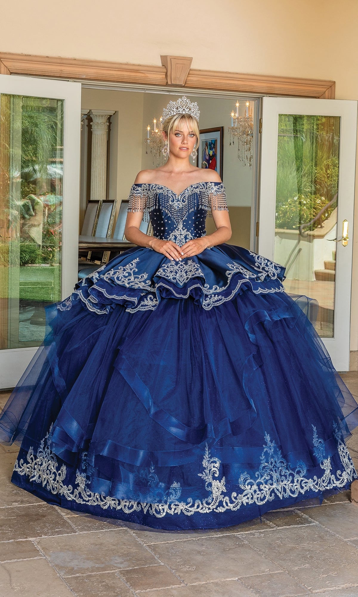A person stands indoors in the Navy Blue Quinceanera Dress 1724 with a sweetheart neckline, silver embroidery, and a tiara, posed before glass doors beneath a sparkling chandelier.