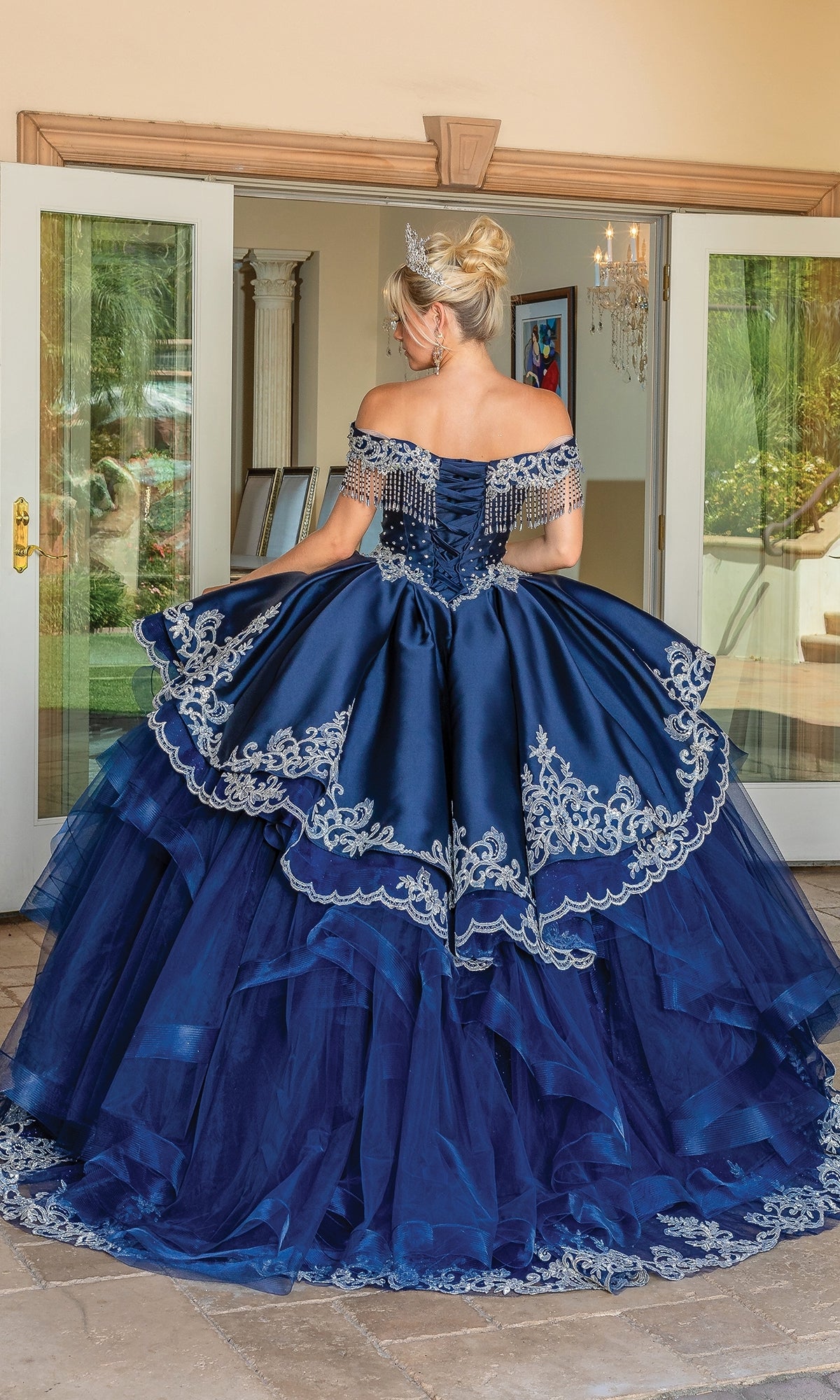 A woman stands indoors facing away, wearing the Navy Blue Quinceanera Dress 1724 with silver embroidery, her hair styled in an elegant updo and topped with a tiara.