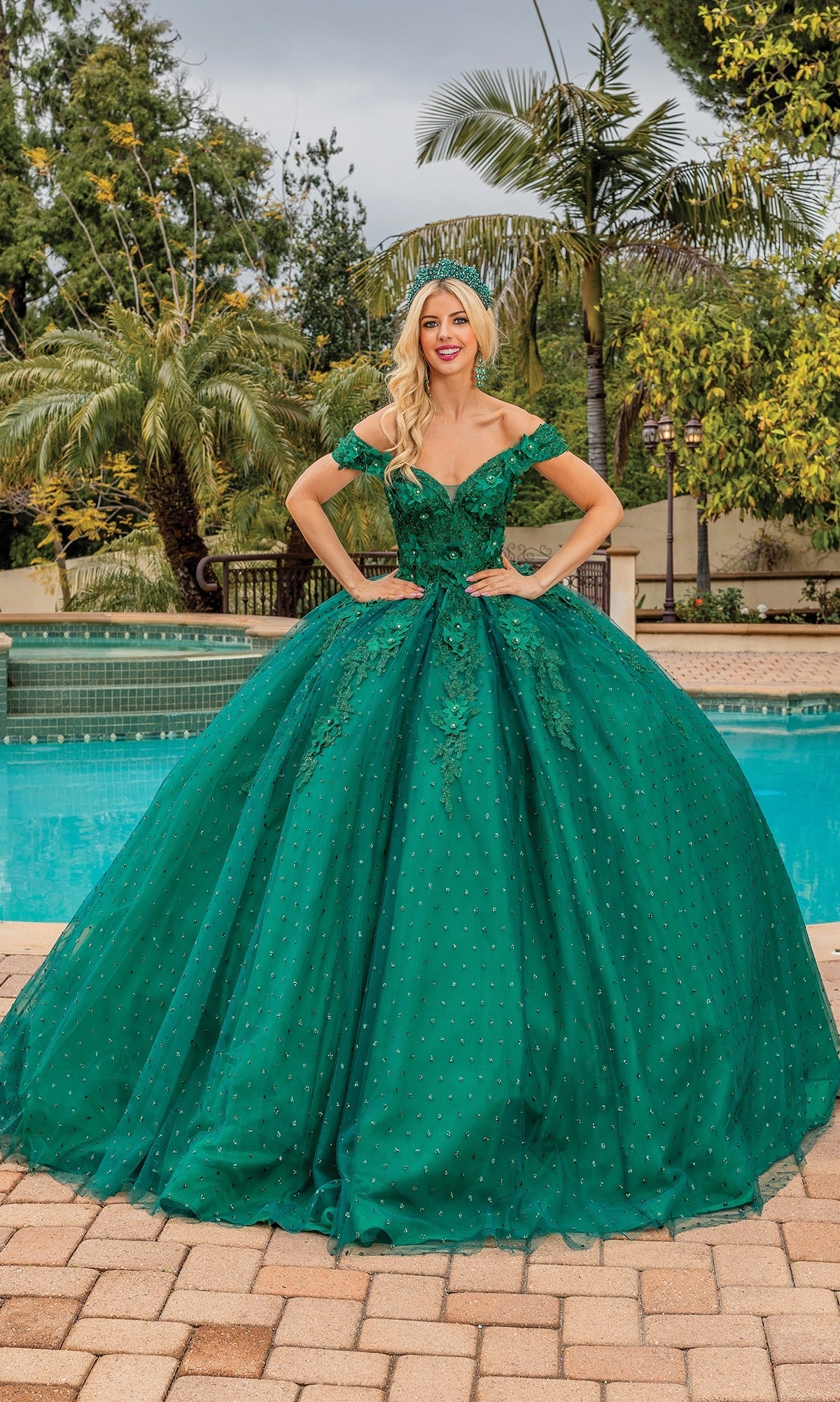 A woman stands outdoors by a pool wearing the Quinceanera Dress 1726 By Dancing Queen, an emerald green off-the-shoulder gown with a full skirt and floral embellishments.