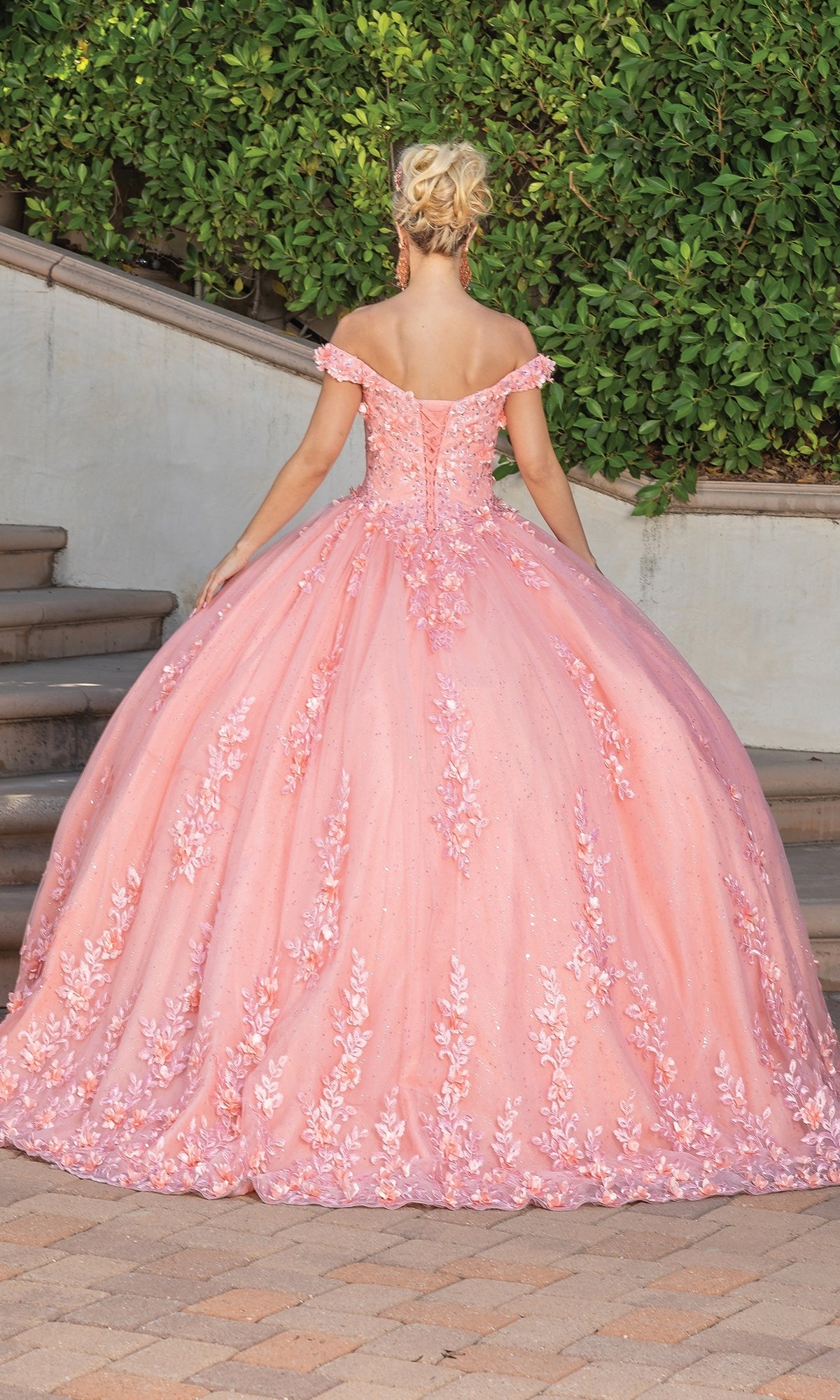 Outdoors, a person faces away from the camera in the Quinceanera Dress 1727 By Dancing Queen, featuring a coral pink ball gown style with floral appliqués and a sweetheart neckline.