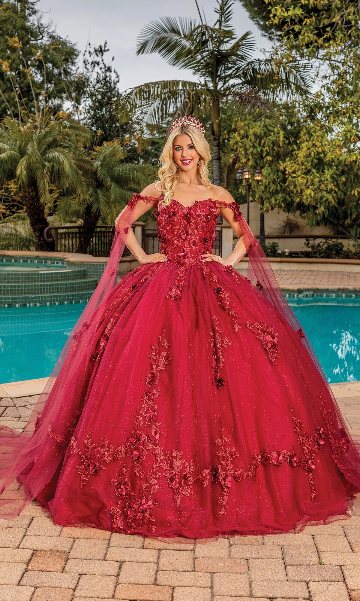 A woman in the Quinceanera Dress 1738 By Dancing Queen with an off-the-shoulder, sweetheart neckline and floral embroidery stands by a pool, surrounded by palm trees and lush greenery.