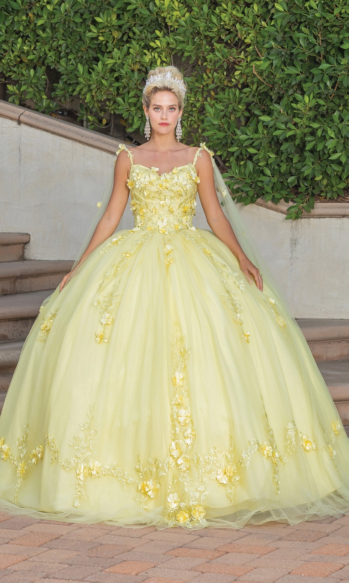 A woman stands outdoors in front of greenery and stone steps, wearing the Quinceanera Dress 1738 By Dancing Queen—an off-the-shoulder pale yellow gown with floral embellishments, paired with earrings and a headpiece.