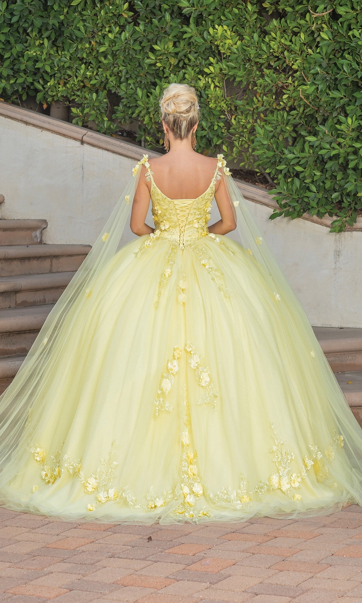 A person with blond hair in an updo wears the Quinceanera Dress 1738 by Dancing Queen, a flowing yellow a-line ball gown with floral details, standing on a brick path near stairs and greenery, facing away from the camera.