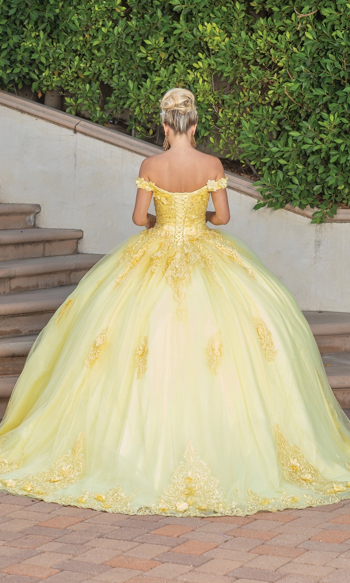 A person with blonde hair in an updo wears the Quinceanera Dress 1739 By Dancing Queen, an off-the-shoulder gown with gold lace embroidery, standing on a brick path near stairs and green bushes, facing away from the camera.