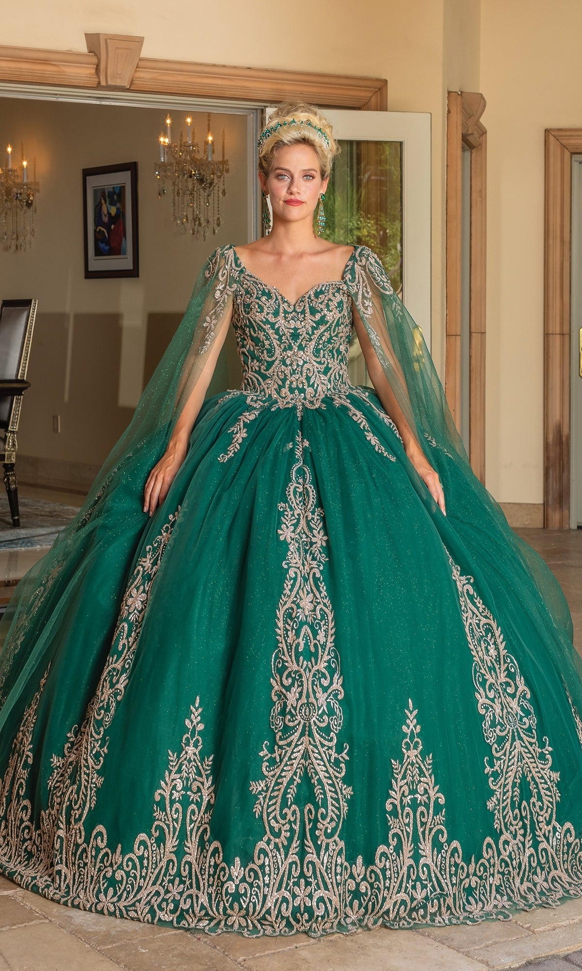 A woman stands indoors wearing the Quinceanera Dress 1743 By Dancing Queen—an emerald green ball gown with gold lace embroidery and draped cape sleeves—looking directly at the camera.