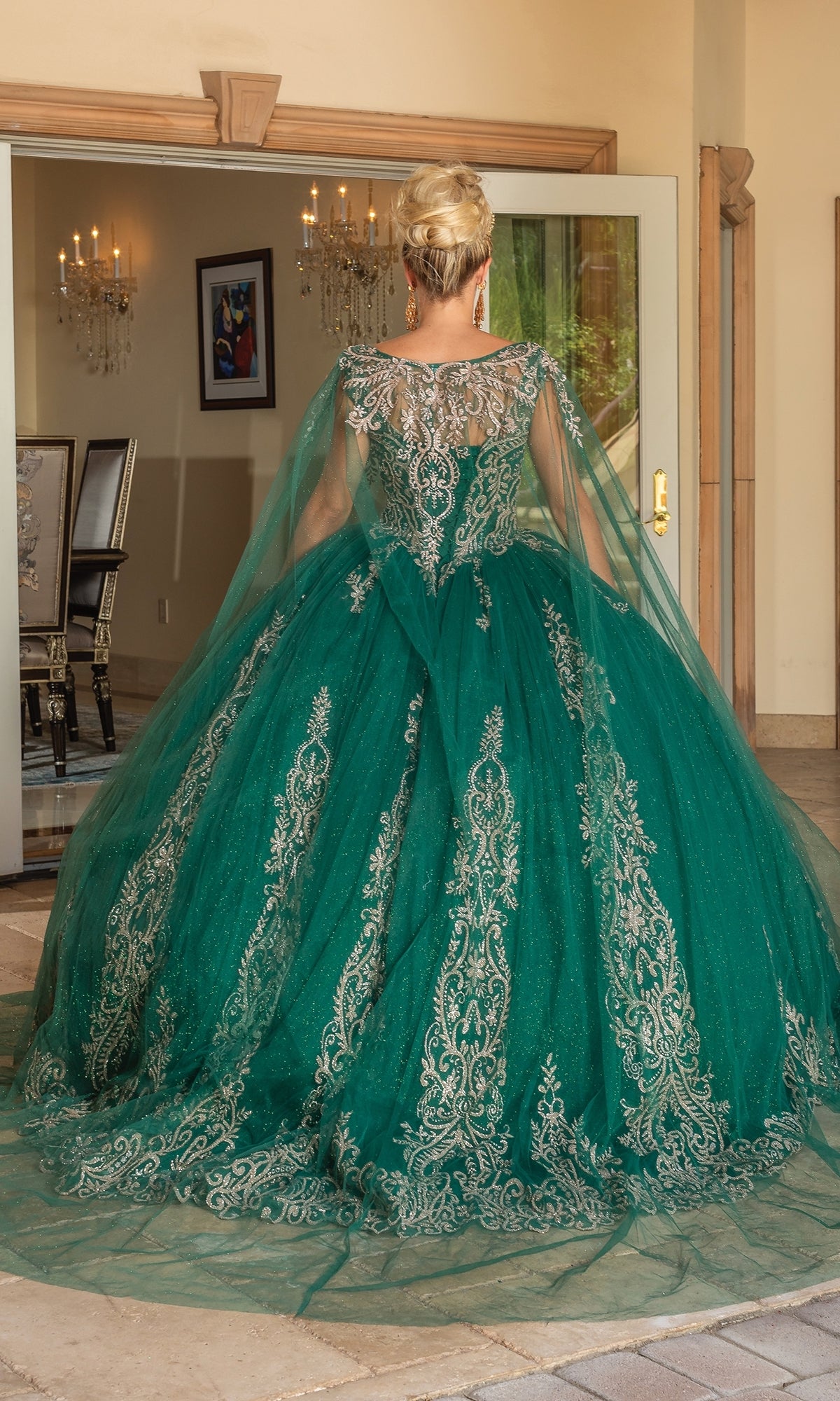 A woman with blonde hair in an updo stands with her back to the camera in front of open glass doors, wearing Quinceanera Dress 1743 By Dancing Queen—an ornate green ball gown featuring gold lace embroidery and a long tulle train.