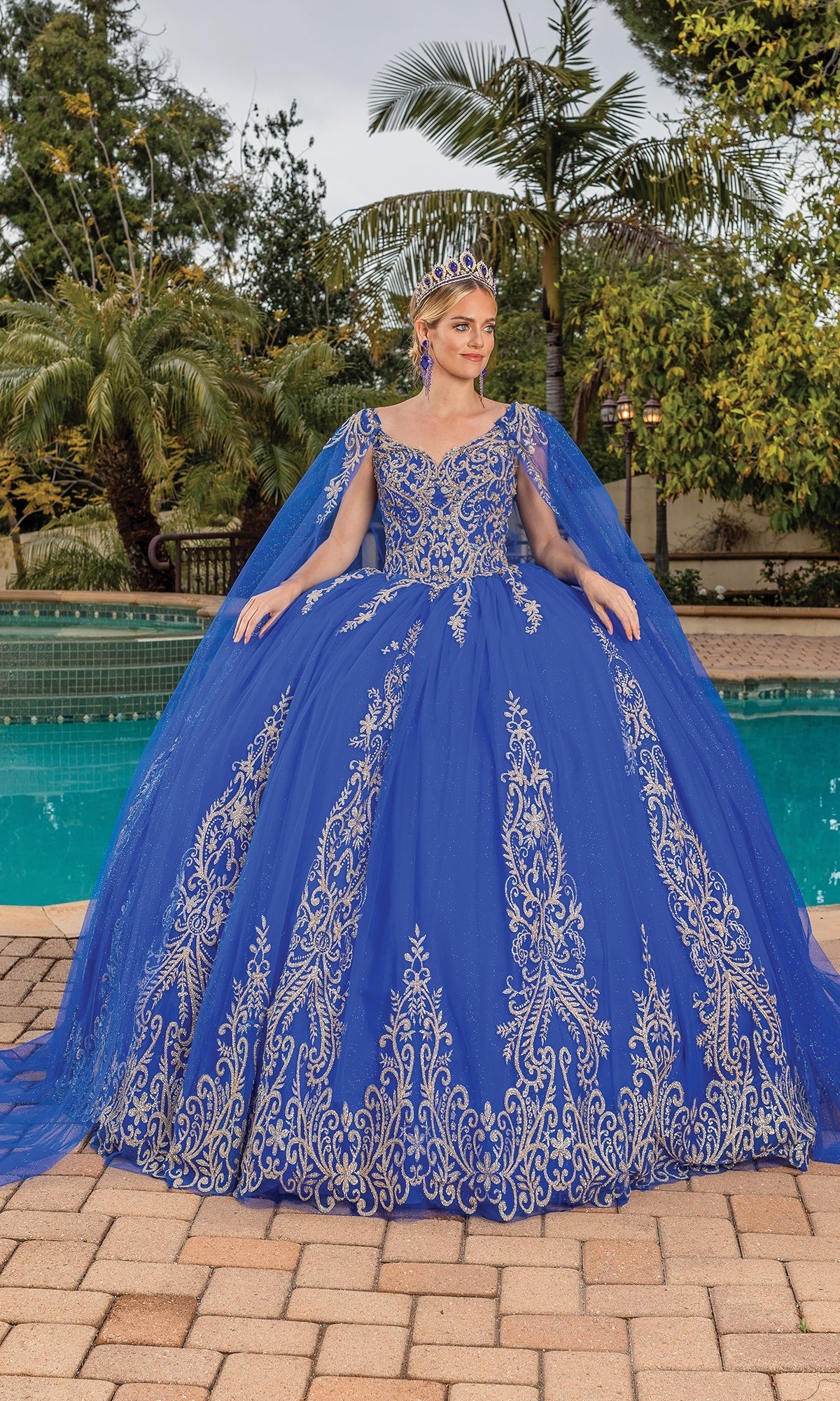 A woman stands by a pool in Quinceanera Dress 1743 By Dancing Queen, a royal blue ball gown with gold lace embroidery and cape sleeves, wearing a tiara and surrounded by palm trees and lush greenery.