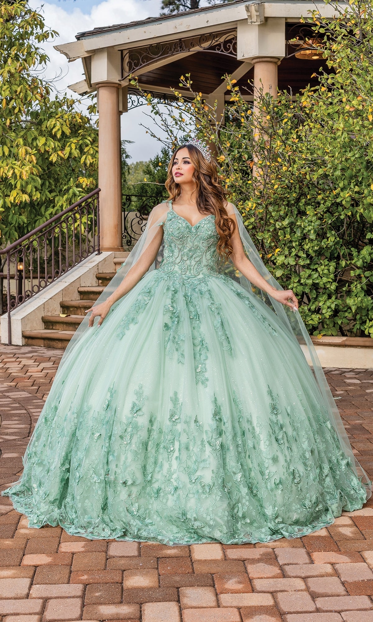 A woman stands on a brick walkway in front of a gazebo, wearing the Quinceanera Dress 1746 by Dancing Queen—a light green, A-line ball gown with floral details, a full skirt, and cape—surrounded by lush greenery.