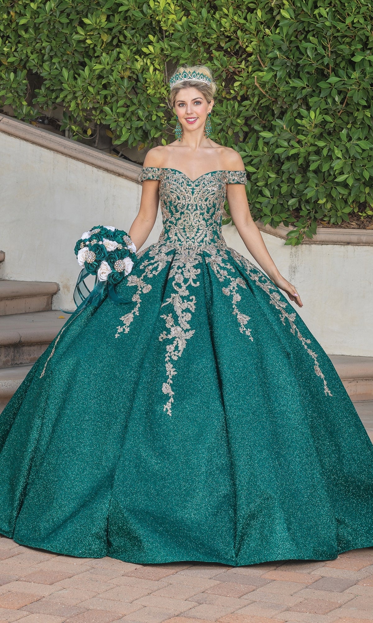 A woman wearing the Quinceanera Dress 1753 By Dancing Queen—an emerald green, off-the-shoulder gown with silver embellishments and lace embroidery—stands outdoors with a matching tiara and bouquet in front of lush greenery and steps.